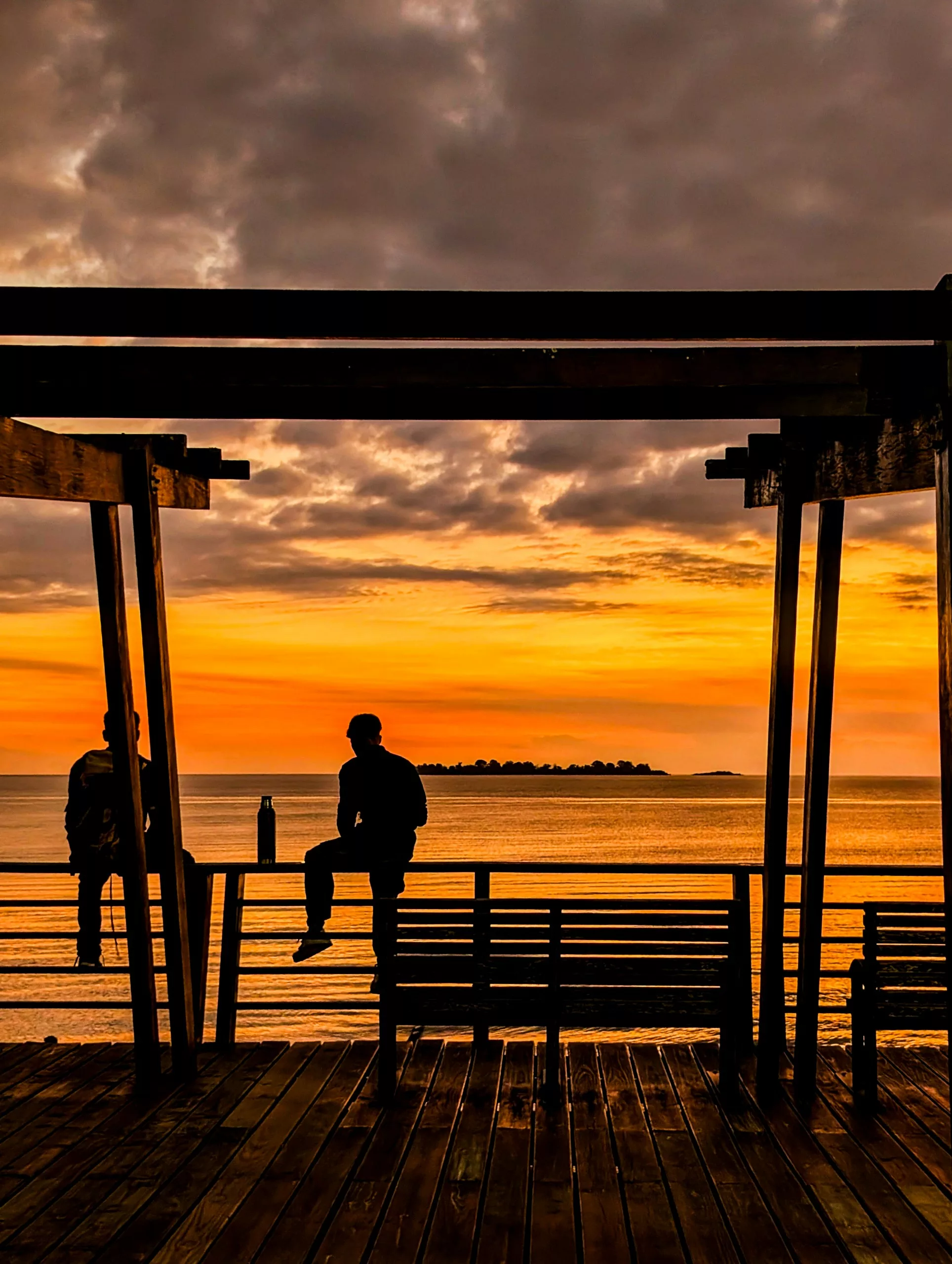 Deux personnes sont assises sur une structure en bois surplombant des eaux calmes au coucher du soleil à Colonia del Sacramento, avec une île au loin et des nuages qui colorent le ciel.