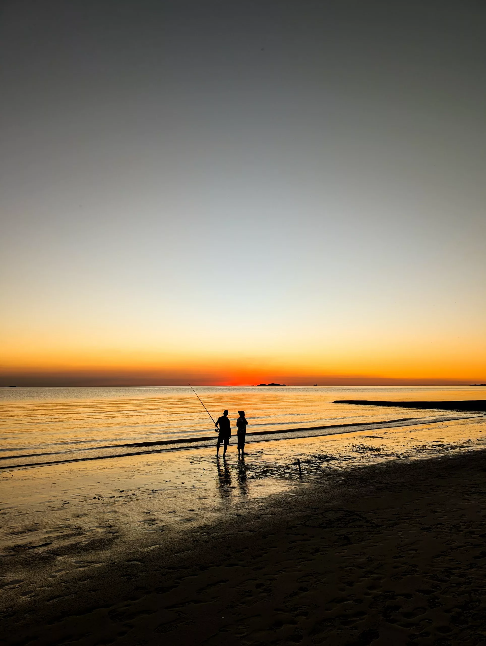 Deux personnes se tiennent sur une plage près du rivage au coucher du soleil à Colonia del Sacramento ; l'une d'elles tient une canne à pêche. Le ciel resplendit de teintes orange et jaunes, qui s'estompent progressivement vers le bleu, tandis que l'océan calme reflète les couleurs du soleil couchant.