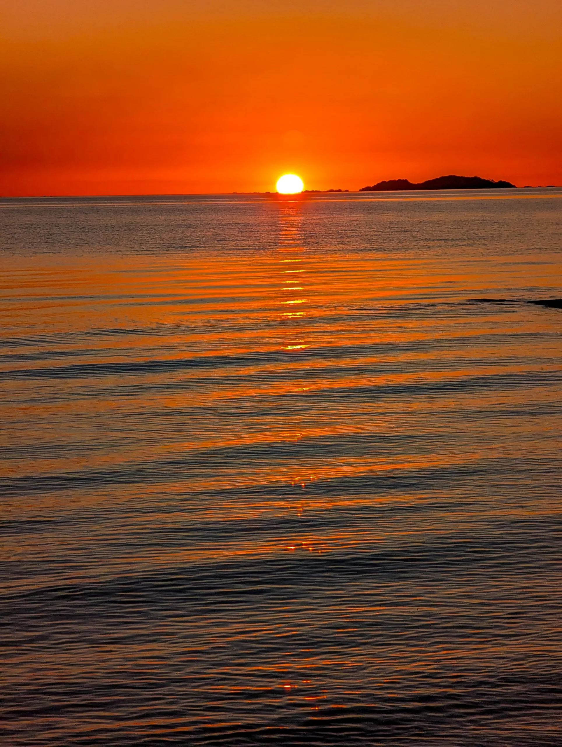 Le soleil se couche sur l'océan près de Colonia del Sacramento, projetant des reflets orange et dorés. Une île apparaît à l'horizon sous un ciel aux couleurs vives, tandis que de douces vagues viennent mourir sur le rivage.