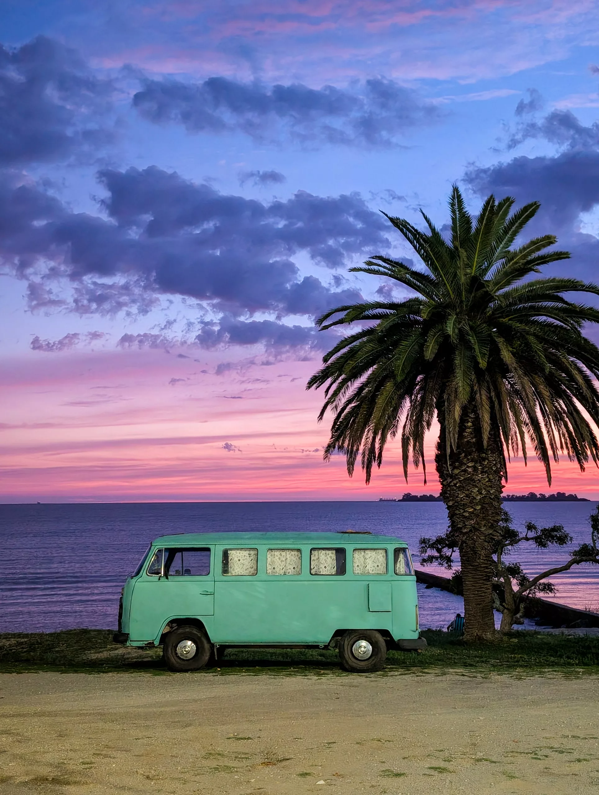 Au coucher du soleil, une camionnette bleu vert est garée près d'un palmier au bord de la mer à Colonia del Sacramento, avec un ciel aux teintes roses et violettes et l'océan en arrière-plan.