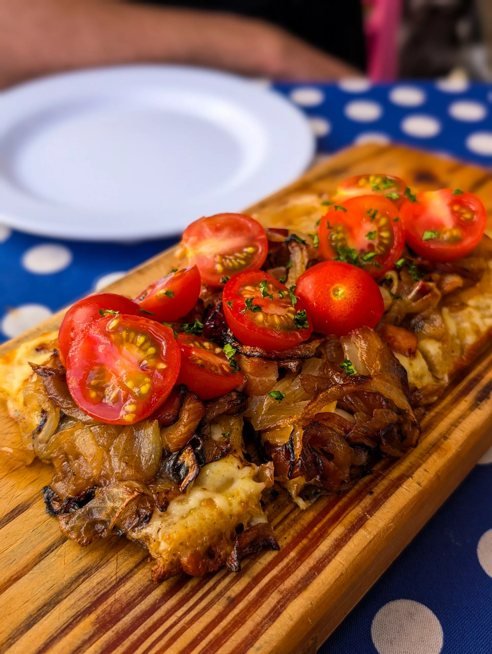 Gros plan sur une planche en bois qui comporte une espèce de grosse tartine recouverte d'oignons frits et de tomates cerises. Le tout repose sur une nappe bleu à pois blanc.
