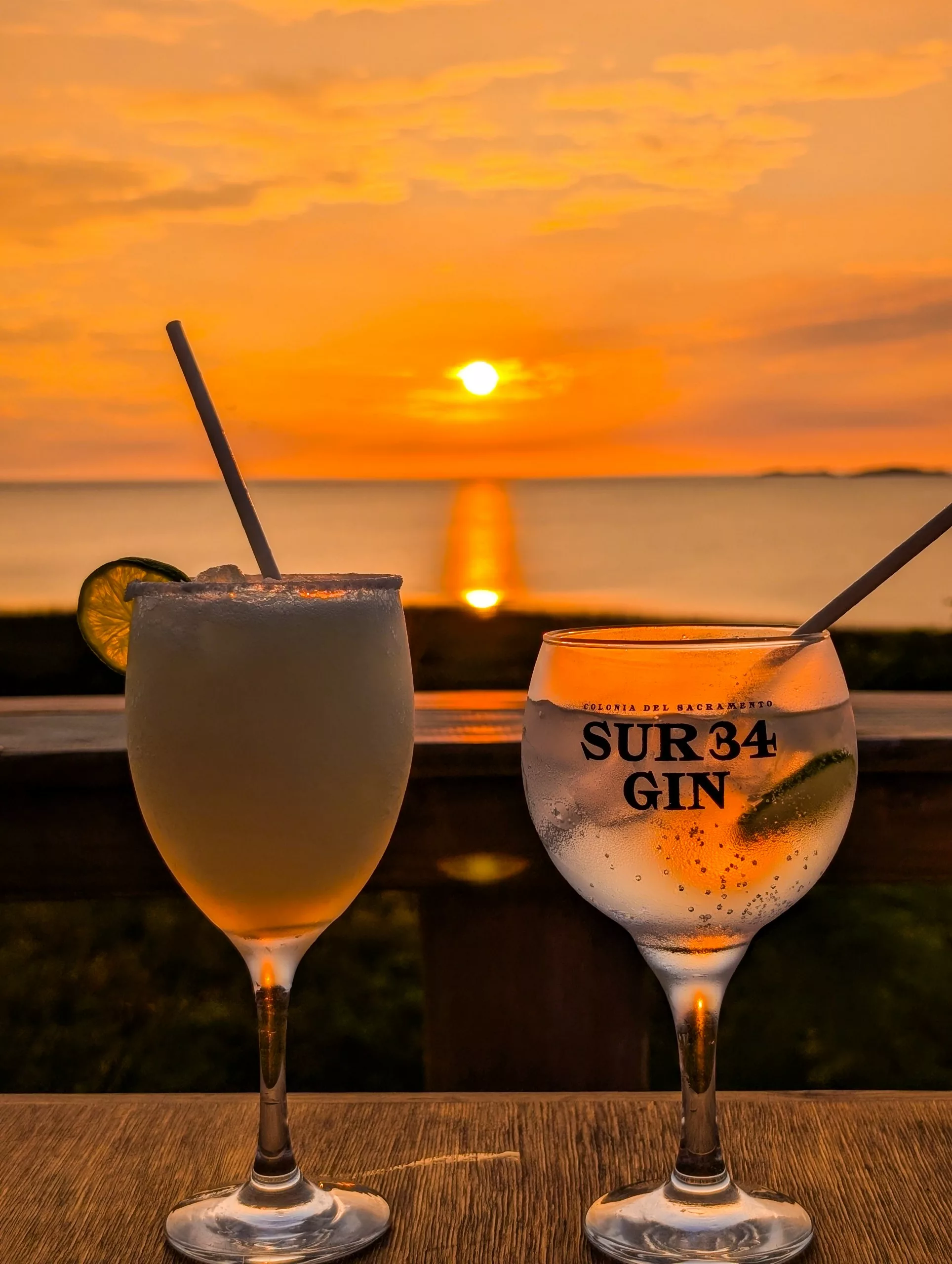 Deux cocktails accompagnés de pailles sont posés sur une table en bois, l'un garni d'un quartier de citron vert, tandis que le soleil se couche sur l'eau à Colonia del Sacramento. Le verre de droite porte l'inscription « SUR 34 GIN ».