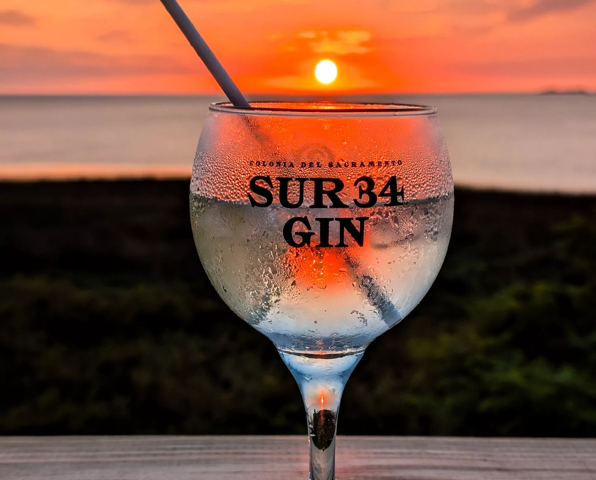 Un verre à gin portant l'inscription « SUR 34 GIN » trône sur une table, accompagné d'une paille et recouvert de condensation, tandis que le soleil se couche sur l'eau, projetant une lueur rouge-orange