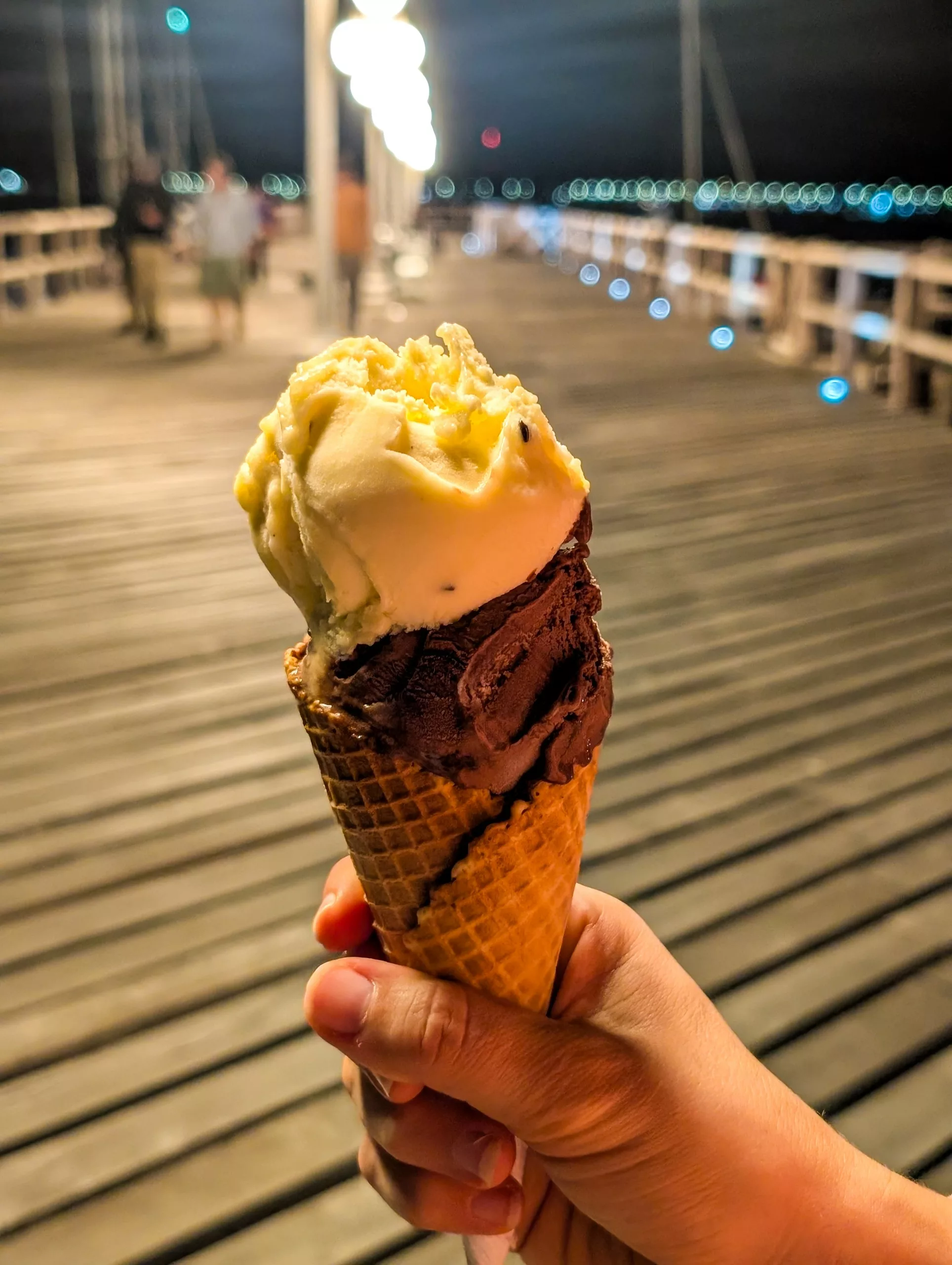 Un cornet de glace avec deux boules : une jaune et une marron. A l'arrière on voit une jetée de nuit, avec des éclairages.