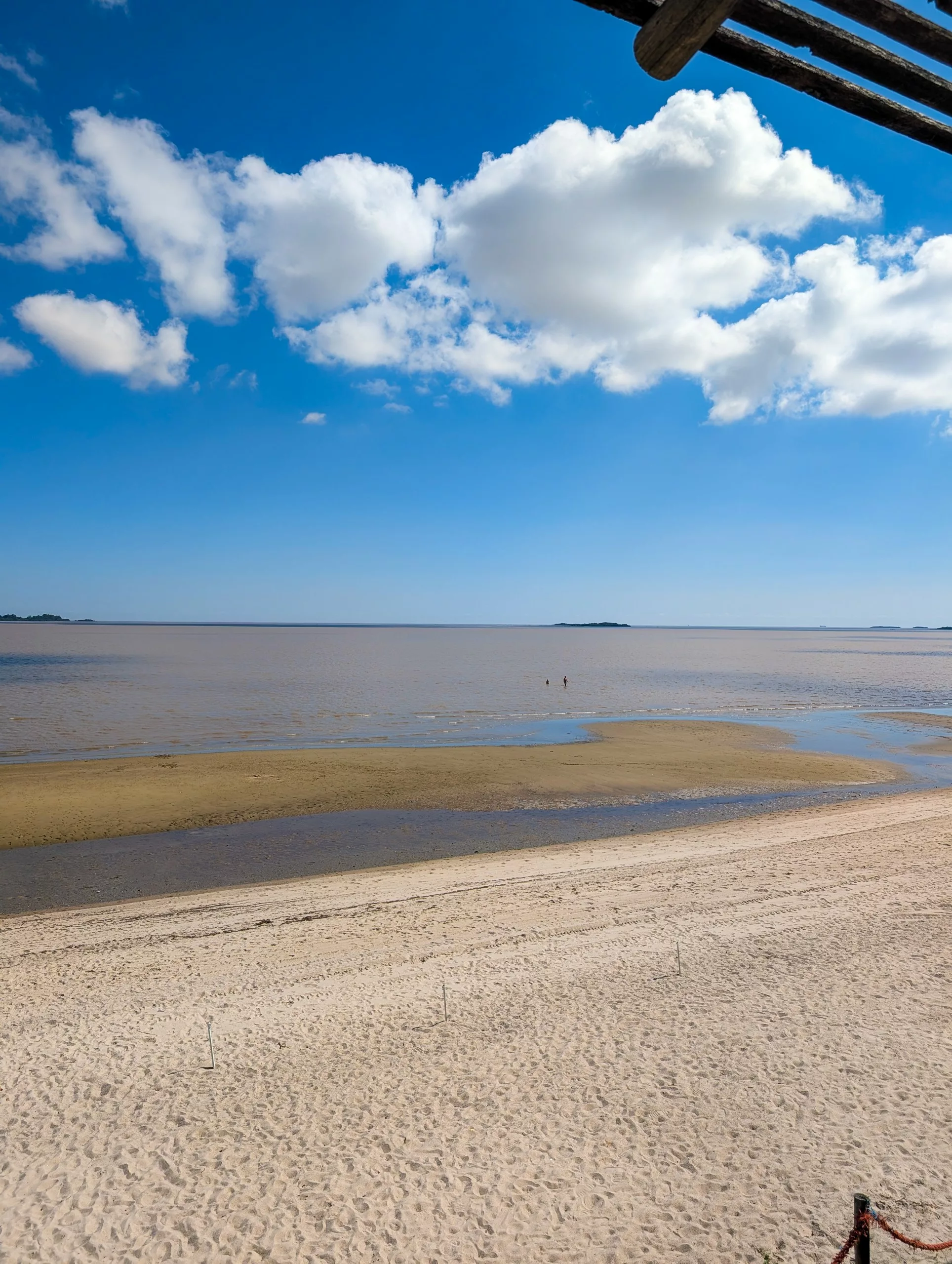 Une grande plage de sable à Colonia del Sacramento mène à des eaux calmes et peu profondes, sous un ciel bleu parsemé de nuages. Deux petites silhouettes lointaines se dressent près de l'horizon, tandis que des îles se profilent au loin, de l'autre côté de l'eau.