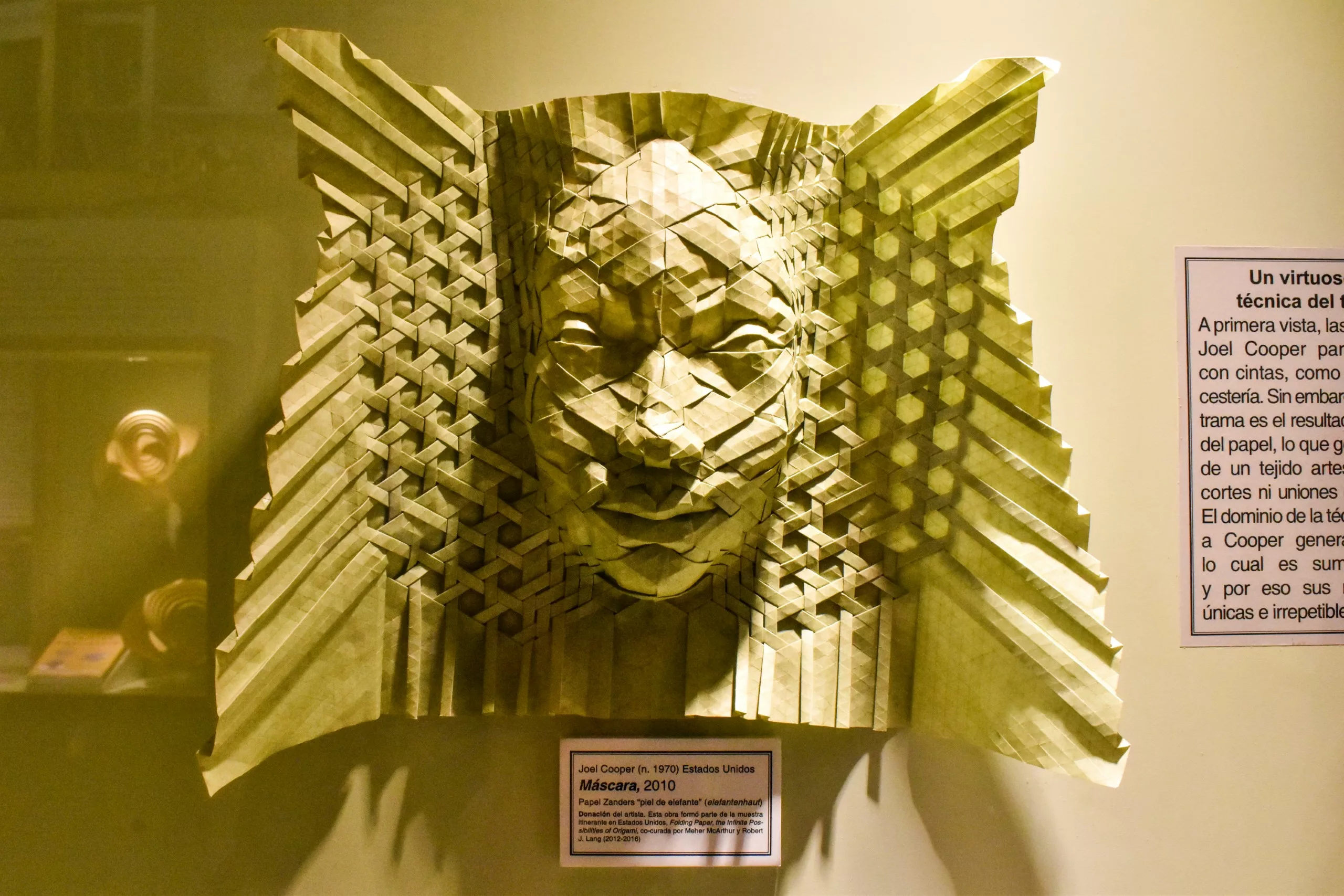 Une sculpture complexe en origami représentant un visage humain est exposée contre un mur ; elle présente un motif géométrique en forme de grille. Une petite plaque informative est visible au pied de l'œuvre.