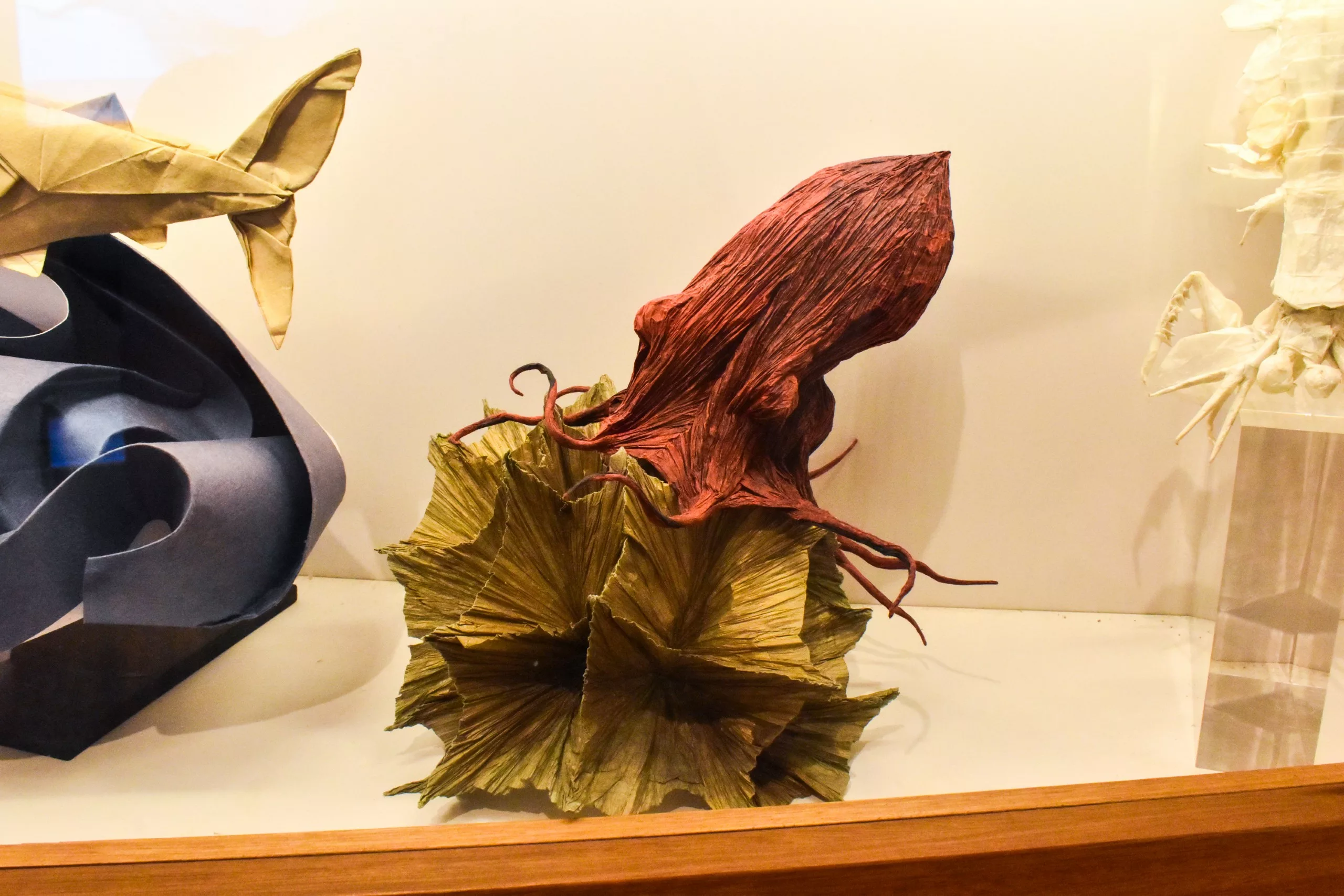 Une sculpture en origami très détaillée représentant un calmar rouge émergeant d'un papier plié vert, exposée dans une vitrine, avec d'autres figures en origami partiellement visibles sur les côtés.