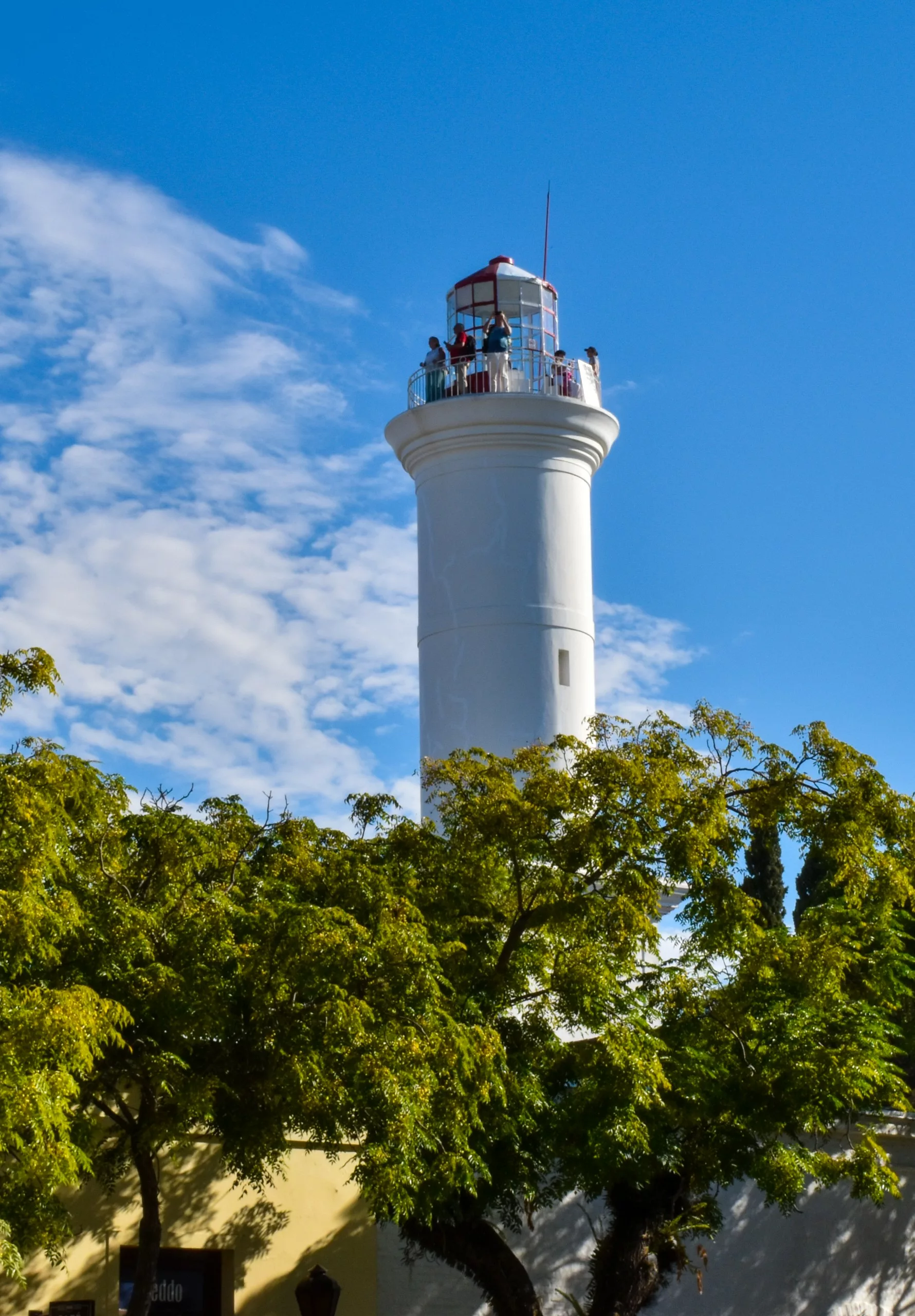 Un grand phare blanc se dresse derrière des arbres au feuillage verdoyant ; on aperçoit plusieurs personnes sur sa plate-forme d'observation. Le ciel est bleu, parsemé de quelques nuages épars.