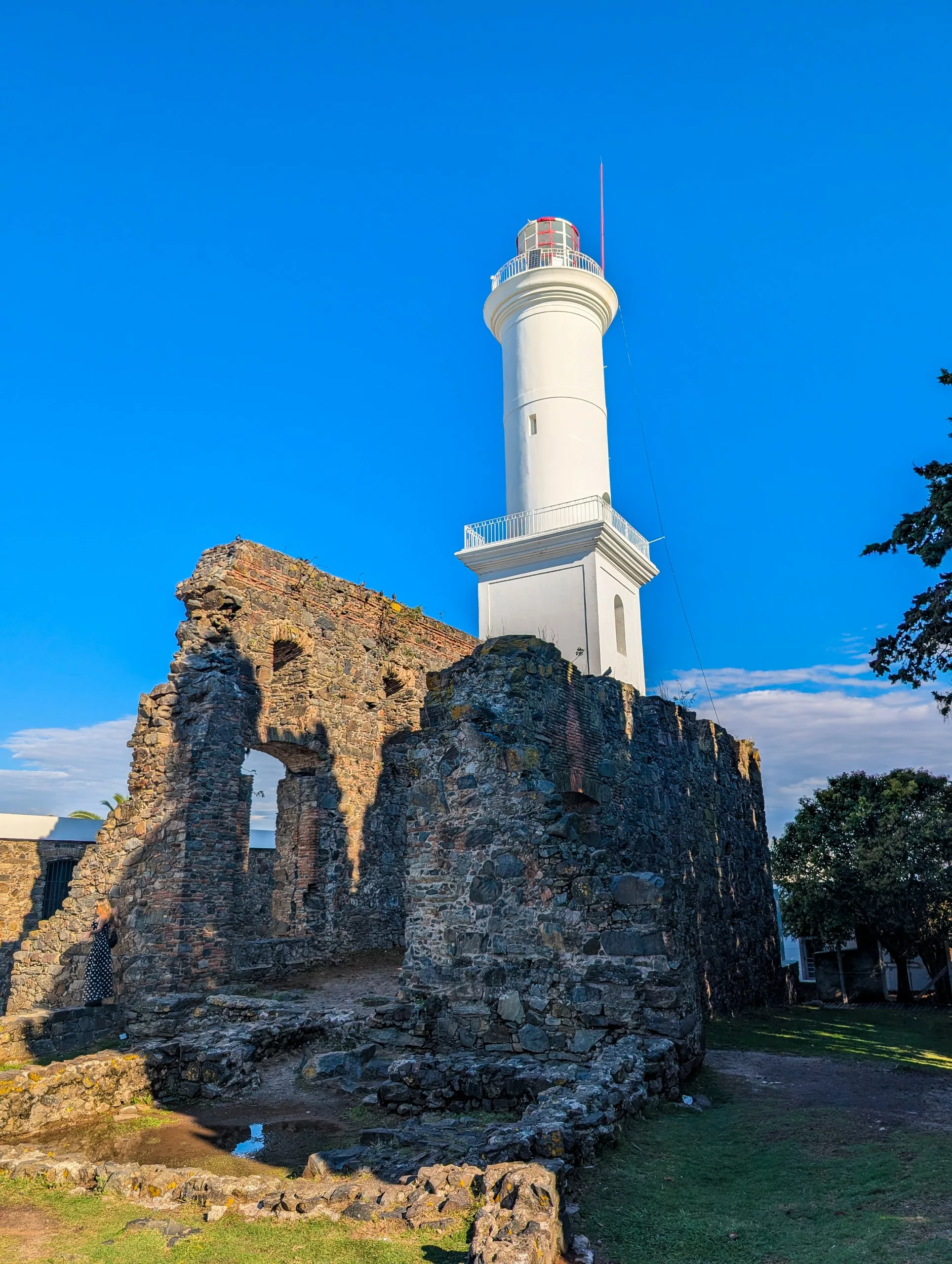 Un phare blanc se détache au-dessus des ruines d'un ancien bâtiment en pierre à Colonia del Sacramento, sous un ciel bleu, avec un premier plan recouvert d'herbe et quelques arbres à droite.
