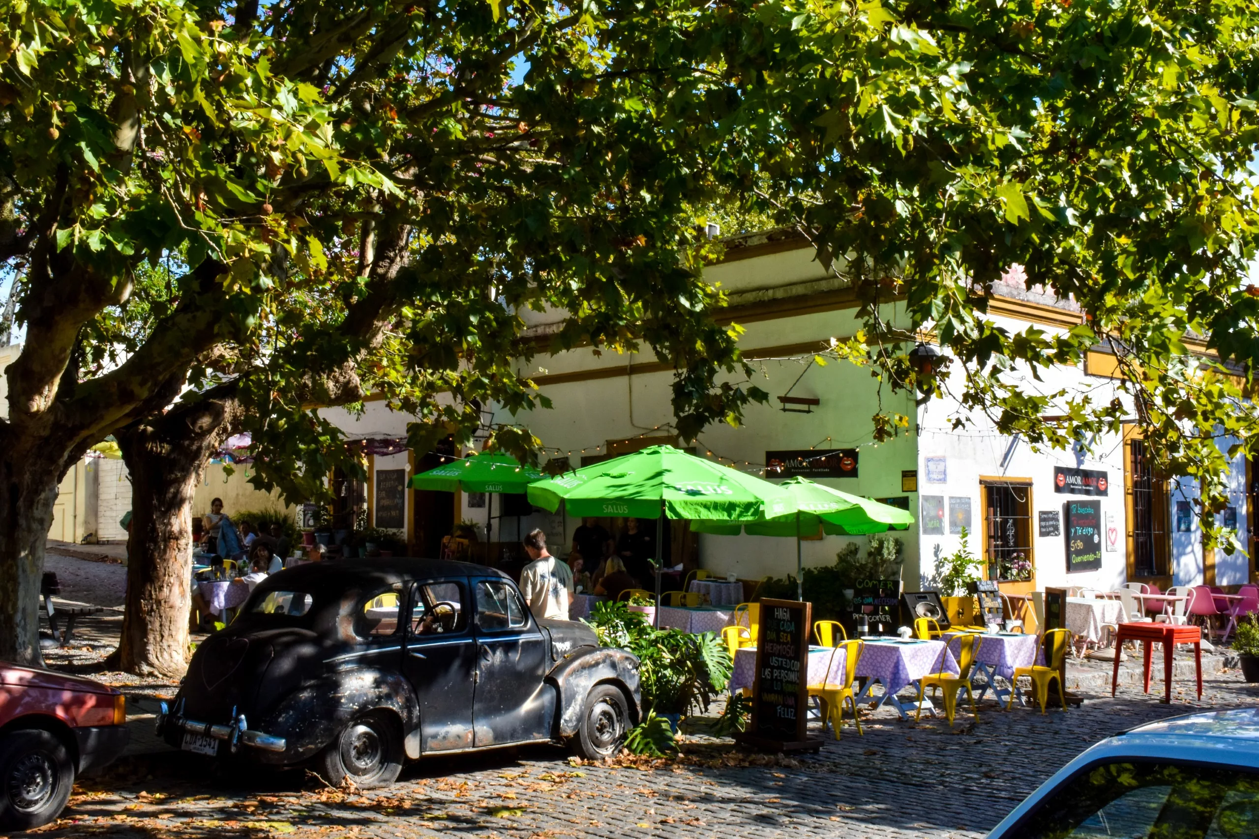 Une vieille voiture noire est garée sous un grand arbre, à côté d'un café aux parasols verts et aux tables colorées, où la lumière du soleil filtrant à travers les feuilles projette des ombres tachetées sur la rue pavée.