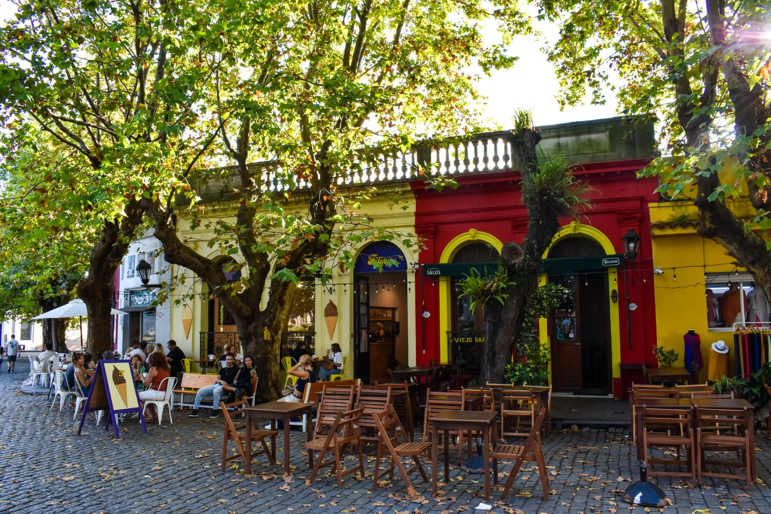 À Colonia del Sacramento, une terrasse de café, avec ses tables et ses chaises en bois vides, se trouve devant des bâtiments aux couleurs vives, jaunes et rouges. Les gens se détendent à l'ombre des arbres , dans une rue pavée.