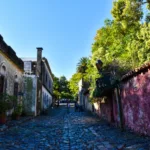 Une rue pavée de Colonia del Sacramento, bordée de part et d'autre de vieux bâtiments patinés par le temps, d'arbres et de verdure, sous un ciel bleu limpide.