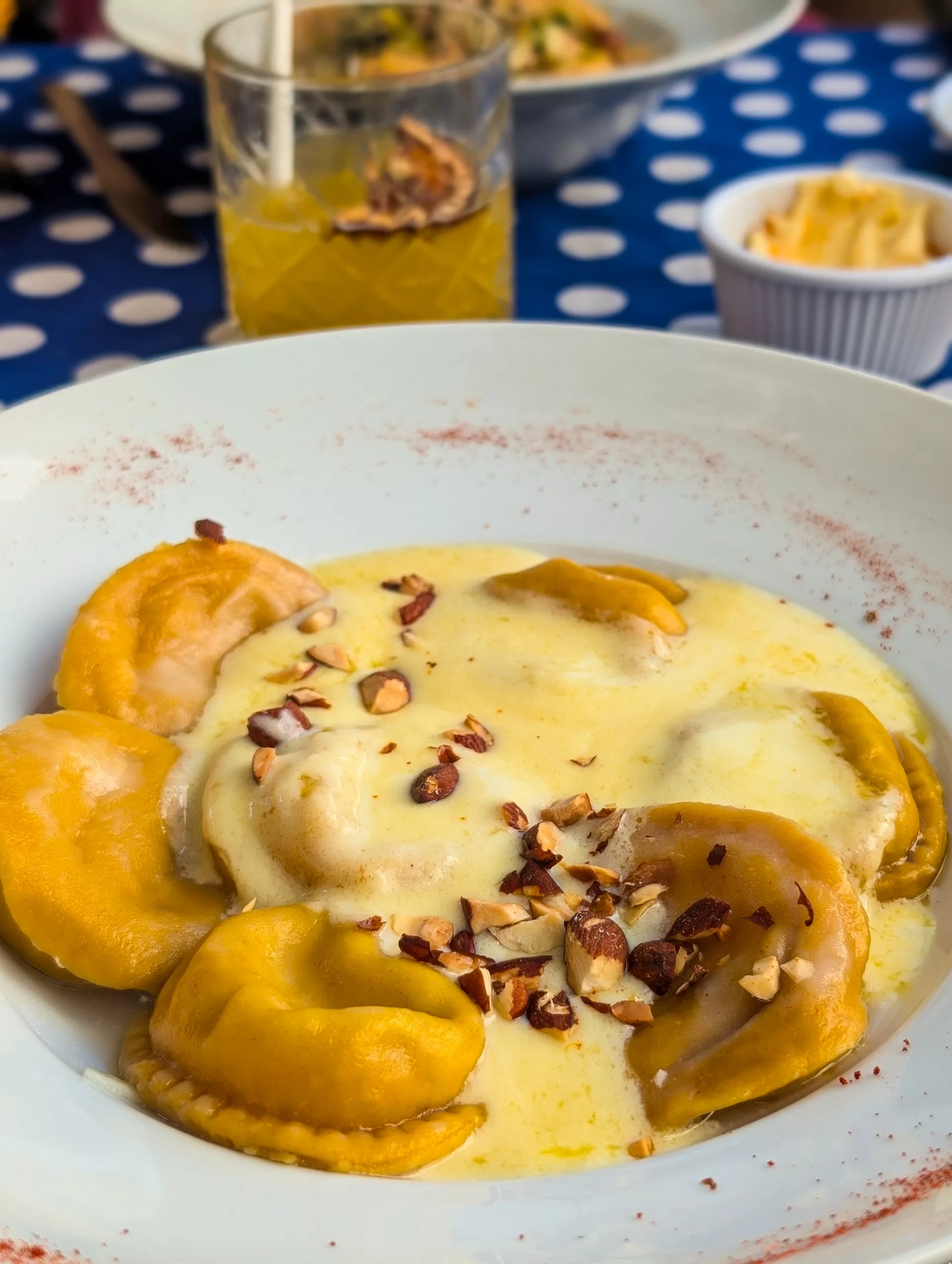 Une assiette blanche de raviolis nappés d'une sauce crémeuse jaune et parsemés de noix hachées, posée sur une nappe à pois bleus et blancs à Colonia del Sacramento. Un verre de cocktail et du fromage râpé sont visibles à l'arrière-plan.
