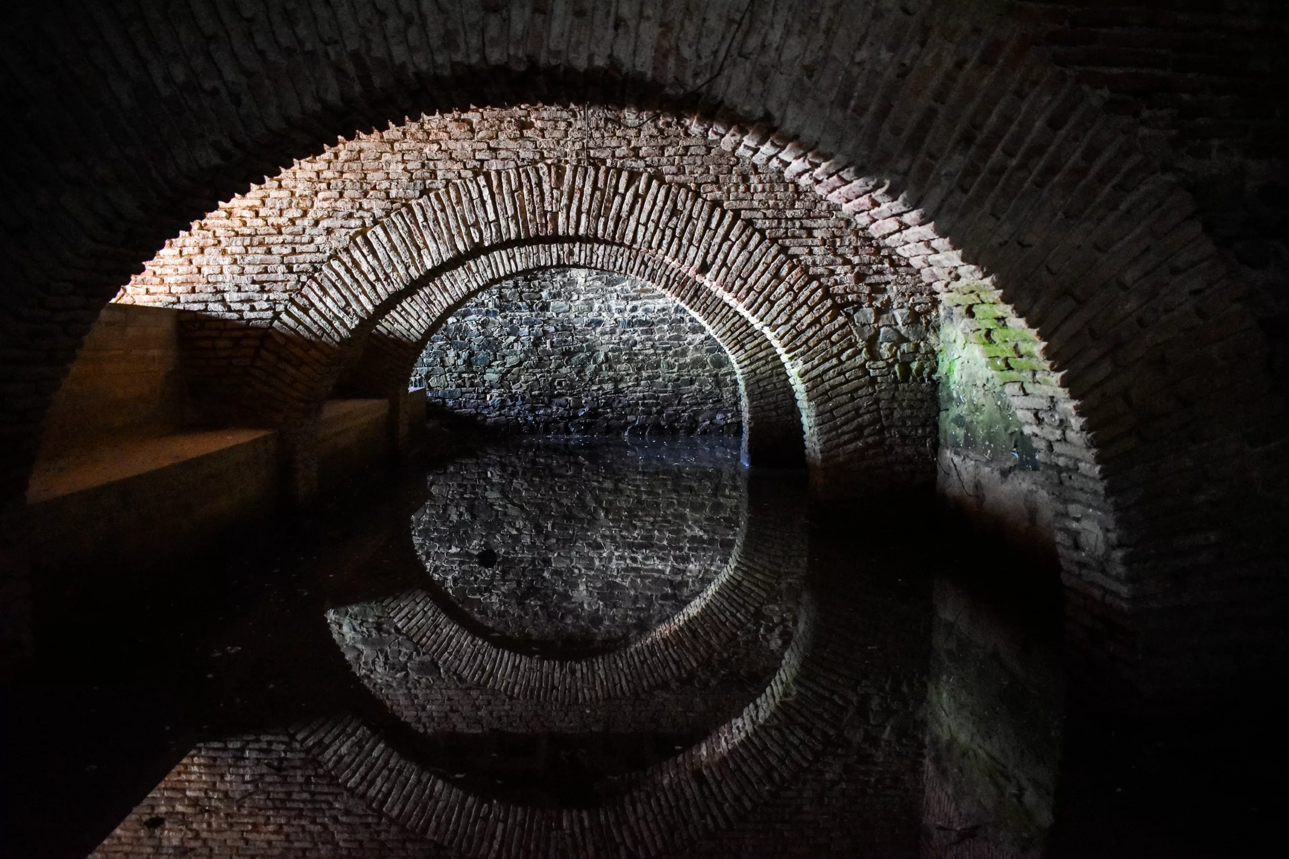 Une arcade en briques située à l'intérieur d'un tunnel sombre se reflète dans l'eau calme en contrebas, formant un cercle presque parfait, tandis que la lumière met en valeur une partie des briques anciennes.