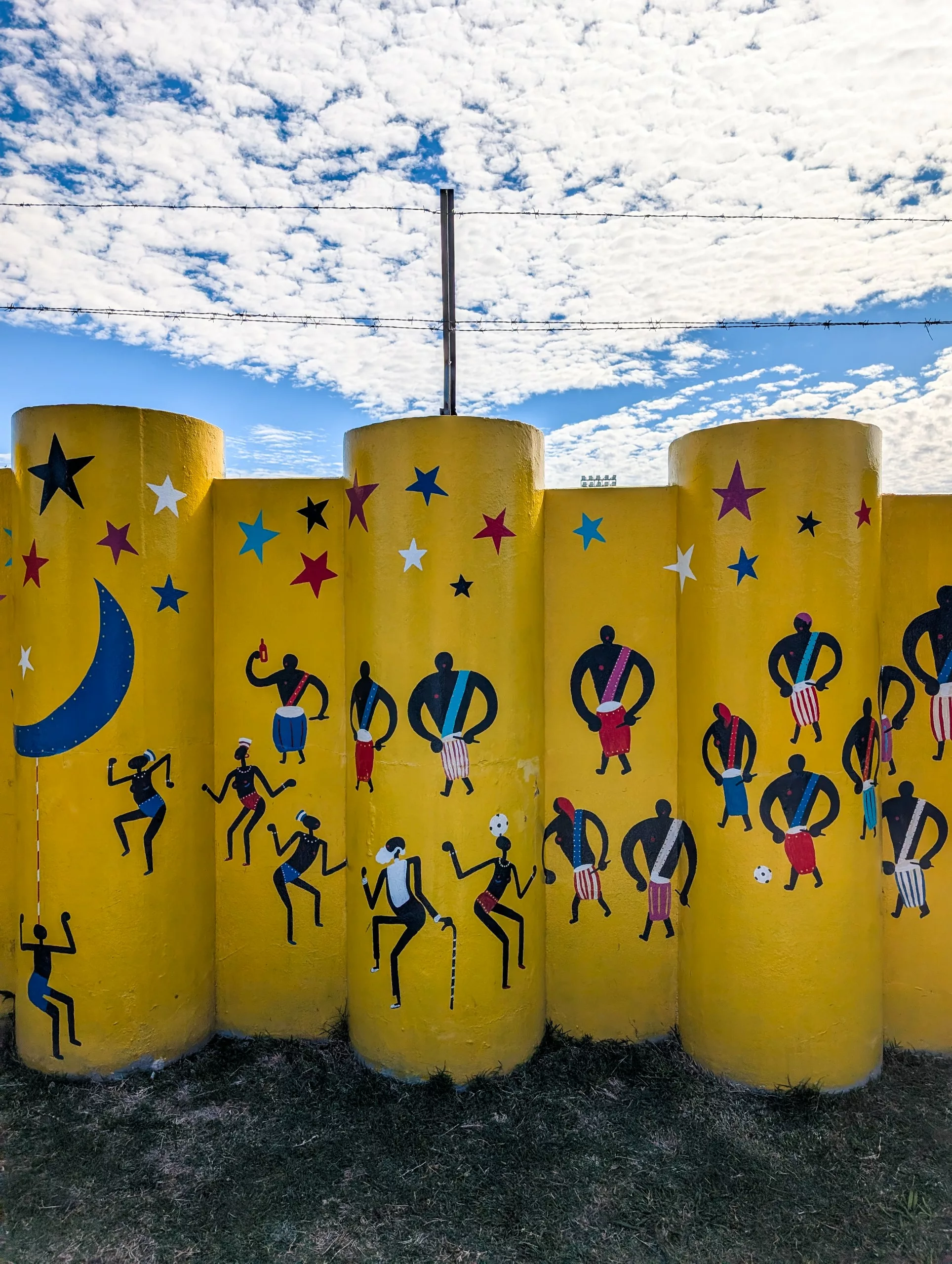 À Colonia del Sacramento, des murs cylindriques d'un jaune vif sont ornés de silhouettes de musiciens, d'étoiles et d'un croissant de lune bleu sous un ciel partiellement nuageux.