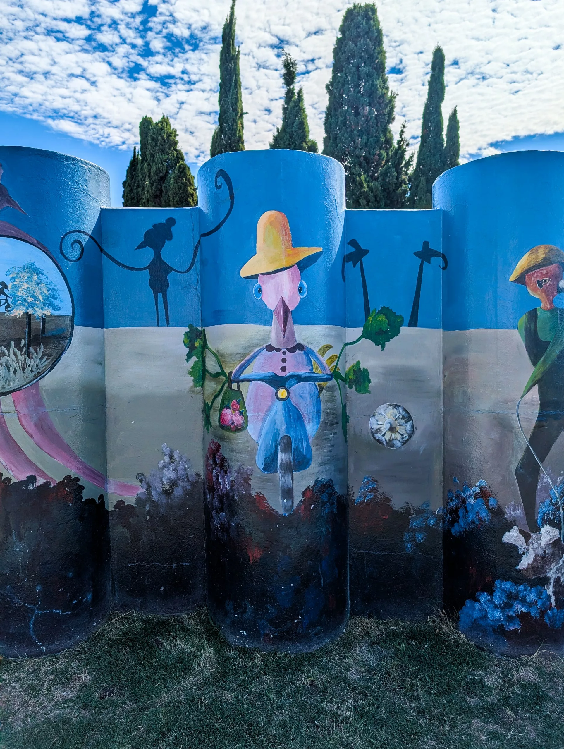 Une fresque murale réalisée sur des piliers bleus arrondis à Colonia del Sacramento représente un personnage central semblable à un oiseau coiffé d'un chapeau jaune, entouré de grappes de raisin et d'un guidon de vélo, flanqué d'autres silhouettes.