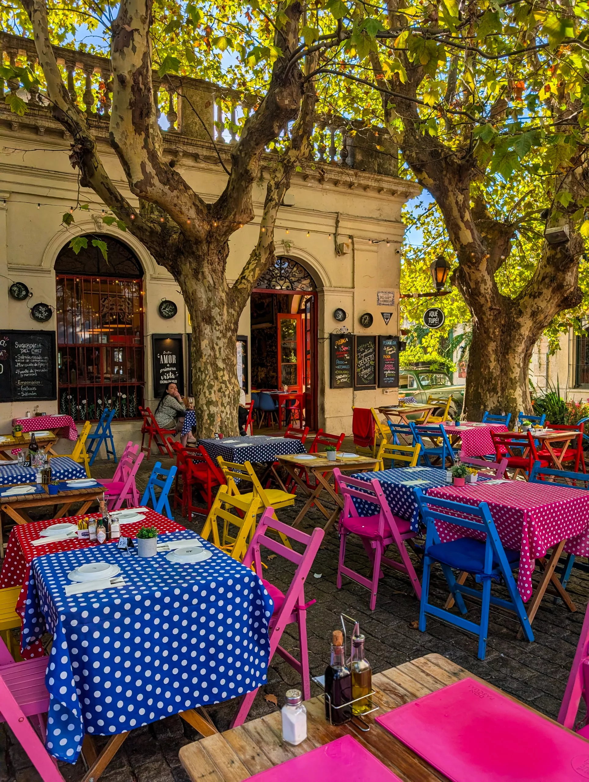 Une terrasse de café à Colonia del Sacramento, avec ses chaises colorées, ses nappes à pois et ses menus écrits à la craie sur des ardoises, à l'ombre d'arbres feuillus, devant un bâtiment beige aux portes cintrées.
