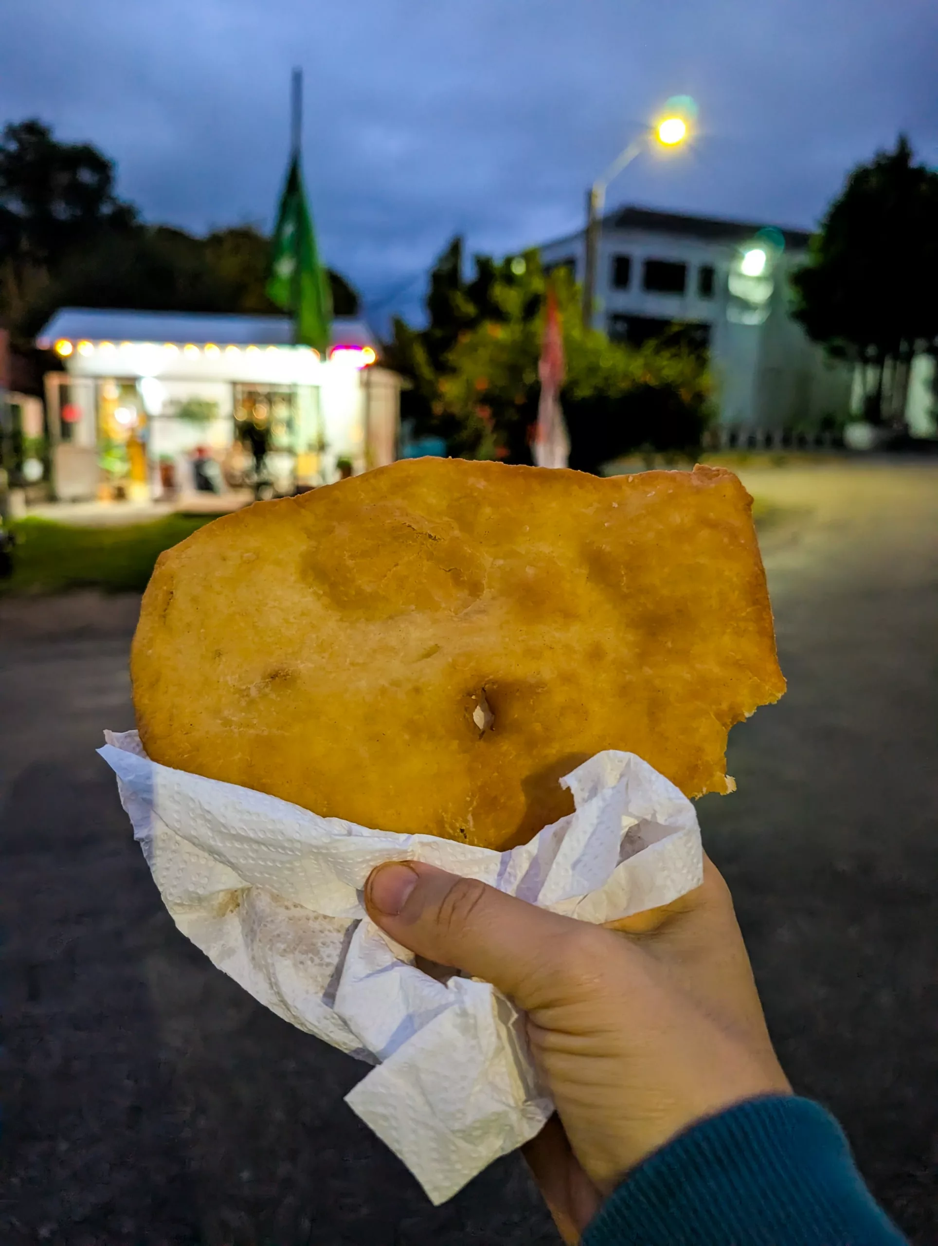 Une main tient un morceau de pâte frite, dont une bouchée a été prélevée, enveloppé dans une serviette. À l'arrière-plan, flou, on aperçoit les lumières d'une rue.