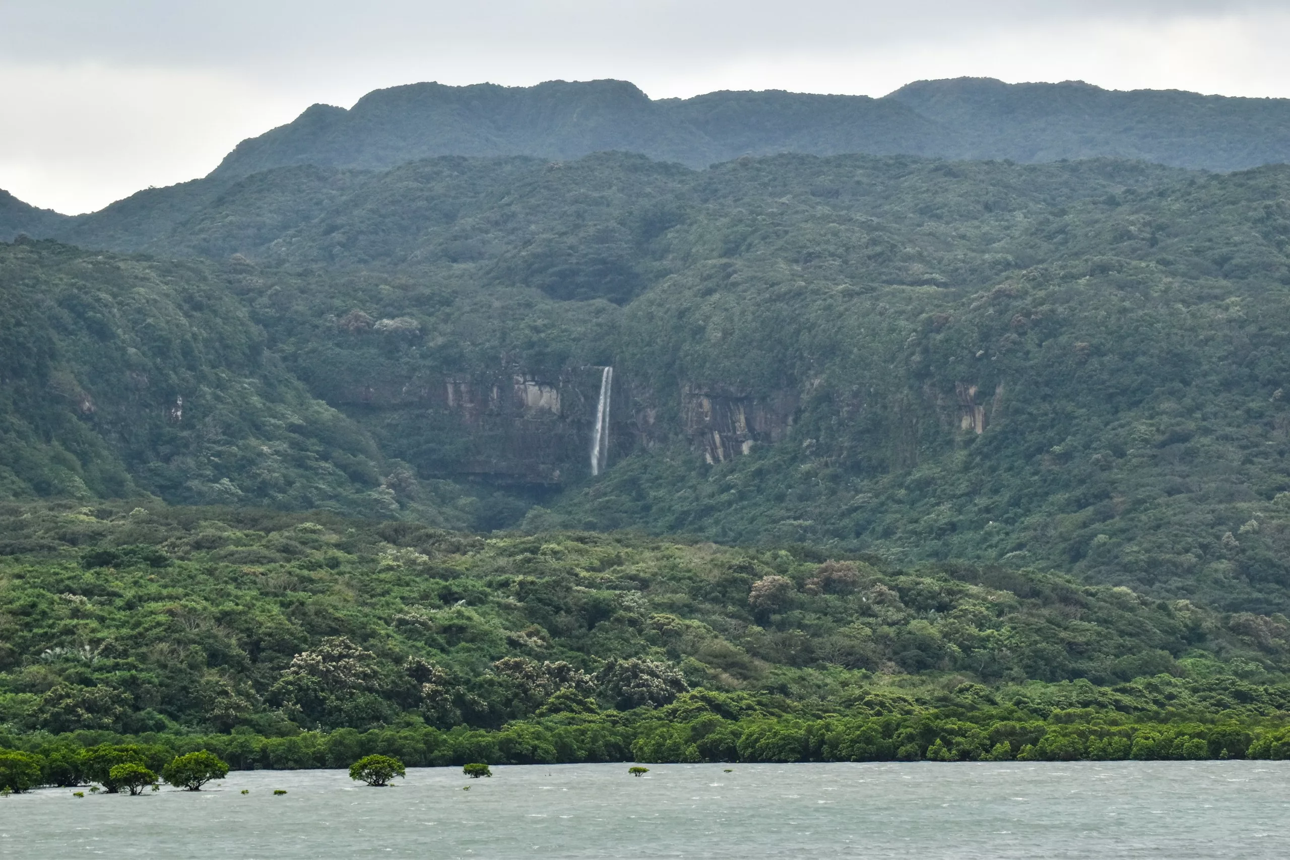 Une haute cascade dévale le flanc d'une montagne boisée au milieu d'une végétation luxuriante, avec un plan d'eau au premier plan et des collines brumeuses sous un ciel nuageux.