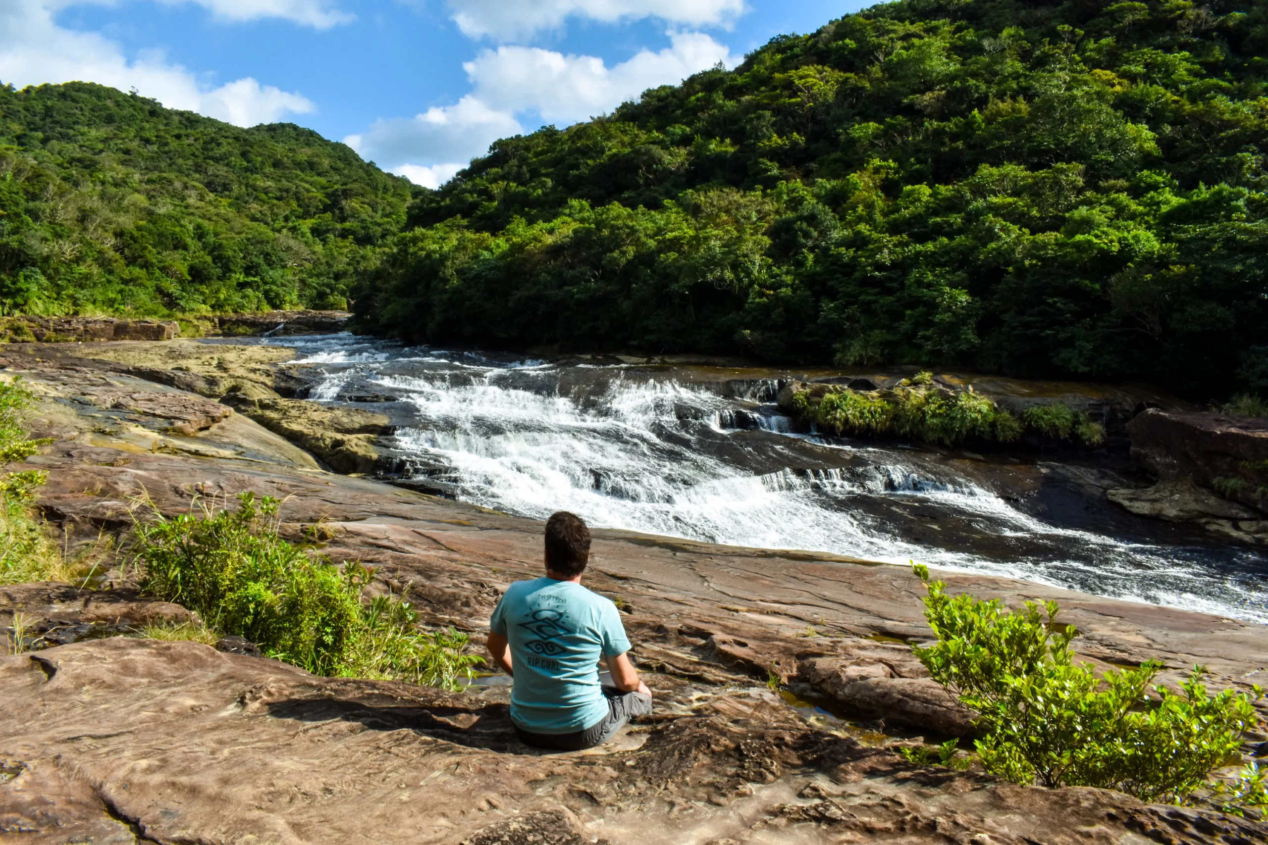 Une personne est assise sur la rive rocheuse d'une rivière à Iriomote, face à une petite cascade entourée d'une forêt verdoyante et dense et de collines luxuriantes, sous un ciel partiellement nuageux.