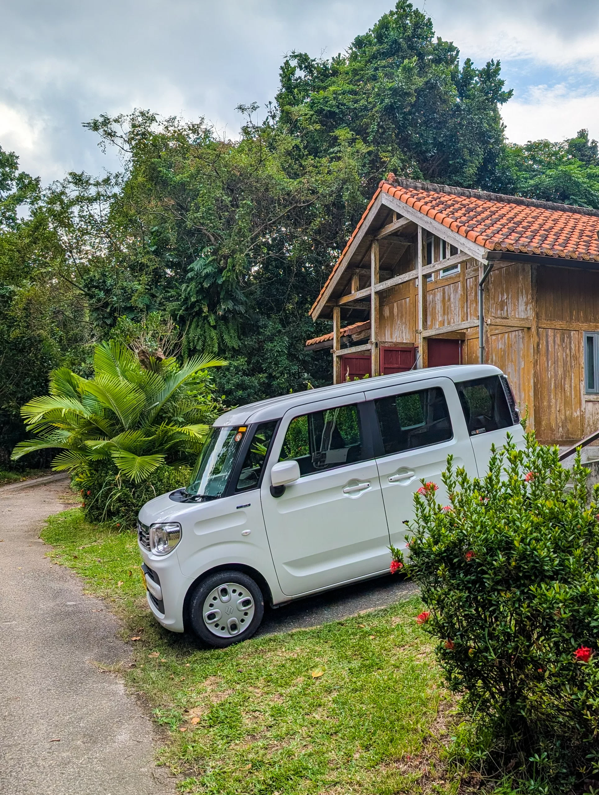 Une voiture blanche japonaise est garée sur la pelouse à côté d'une maison rustique en bois au toit de tuiles rouges, entourée d'arbres et d'arbustes à la végétation luxuriante
