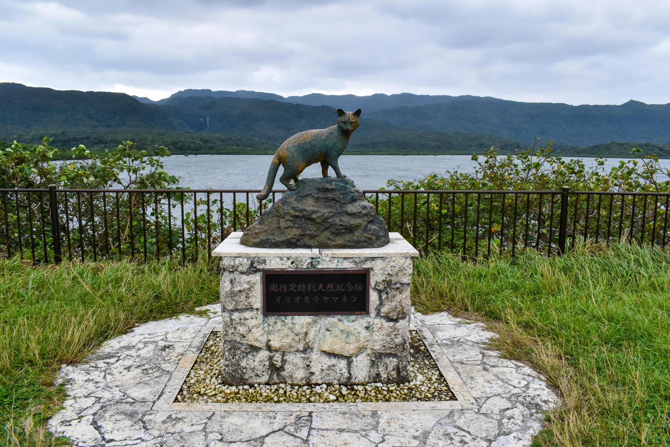 Une statue de chat sauvage en bronze, se dresse sur un socle en pierre au bord de l'eau, entourée de verdure et de montagnes. Une plaque en japonais est fixée sur le devant.