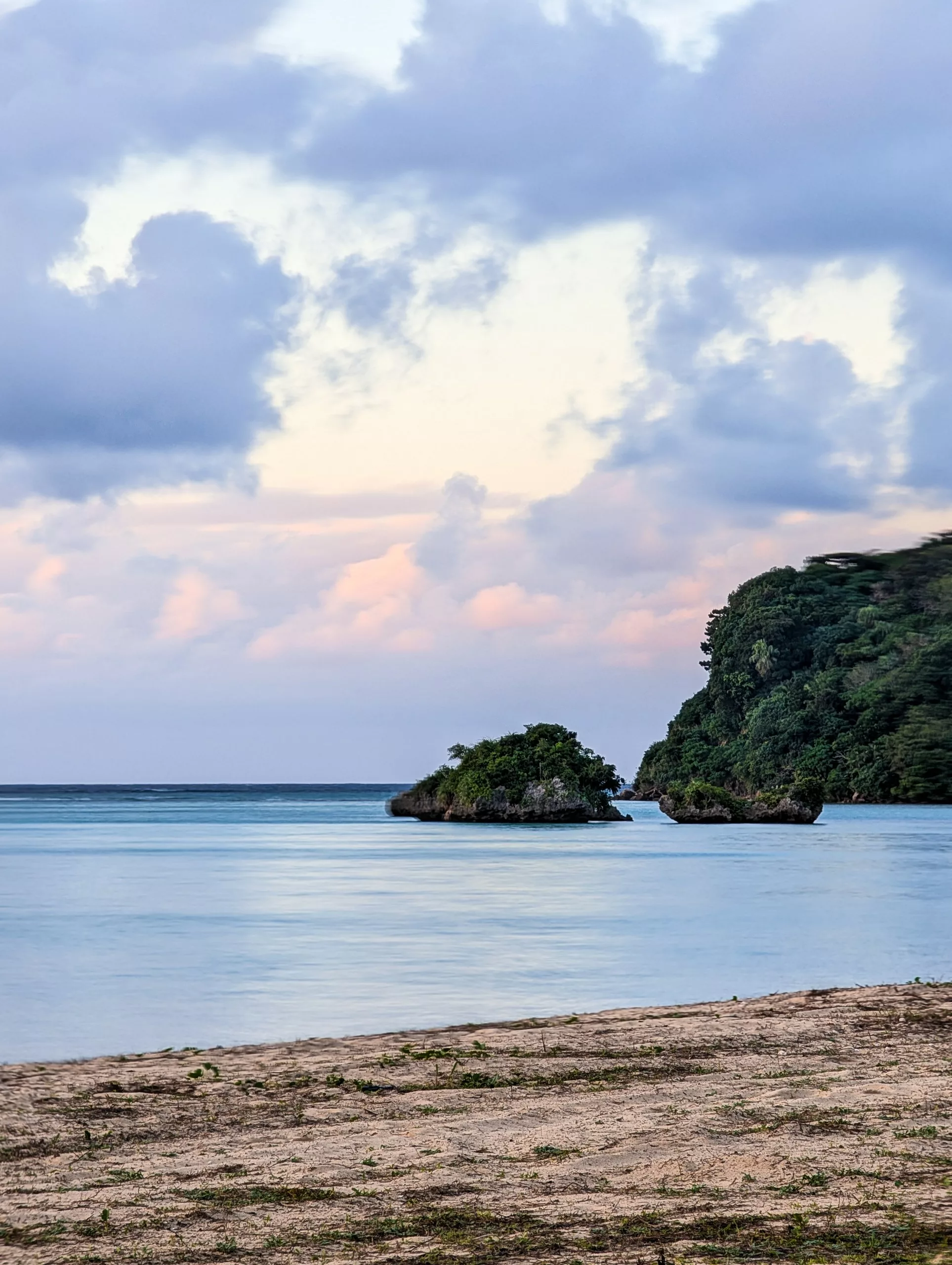 Une scène marine paisible au coucher du soleil avec deux petits îlots rocheux recouverts de végétation, une falaise boisée à droite et une plage de sable au premier plan, sous un ciel nuageux.