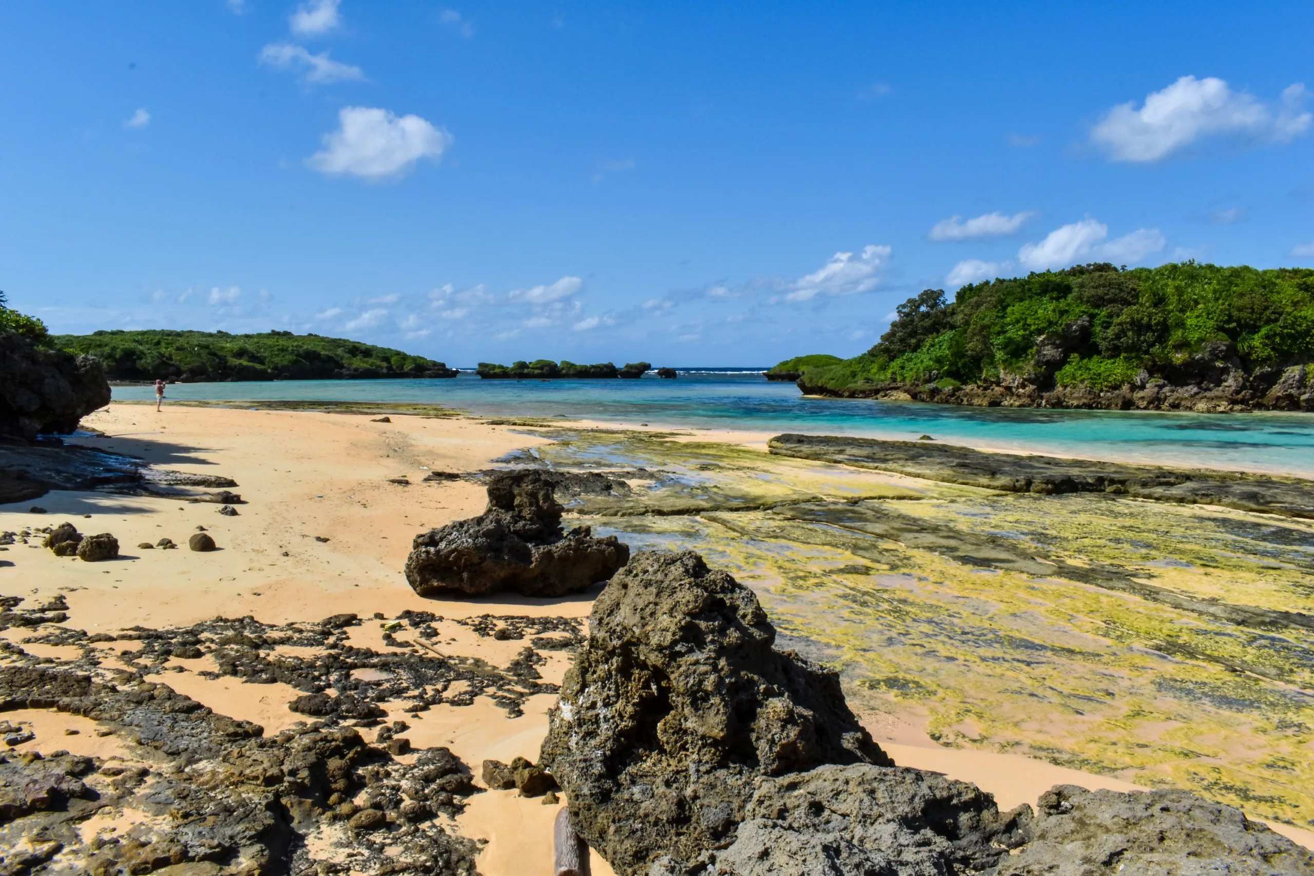 Une plage de sable parsemée de gros rochers et bordée d'une végétation verdoyante. On aperçoit les eaux bleues peu profondes, et une personne marche au loin sous un ciel en grande partie dégagé et ensoleillé.