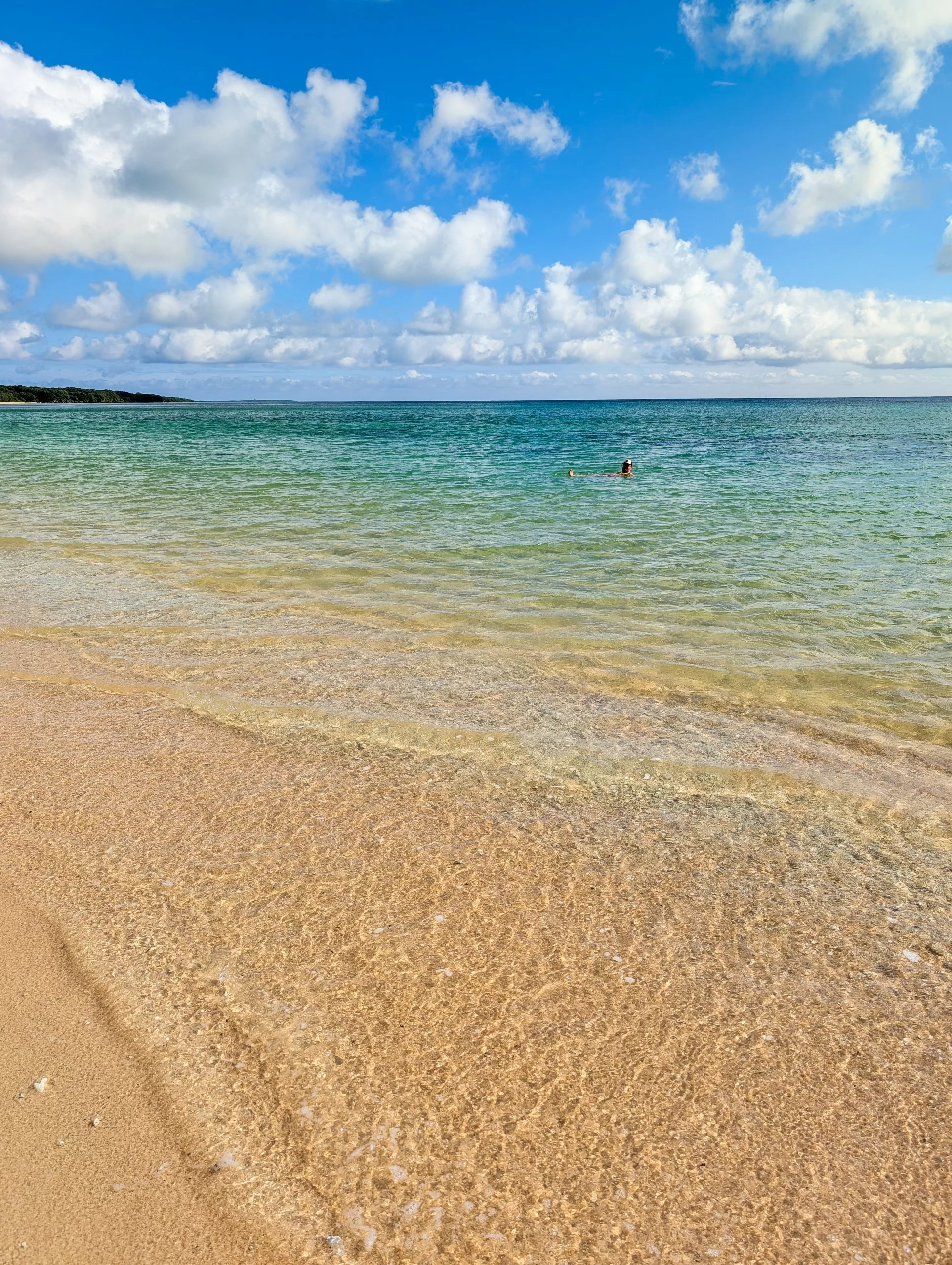Au premier plan, une plage de sable d'Iriomote aux eaux claires et peu profondes. L'océan s'étend jusqu'à l'horizon sous un ciel bleu parsemé de nuages. Au loin, une personne se baigne.