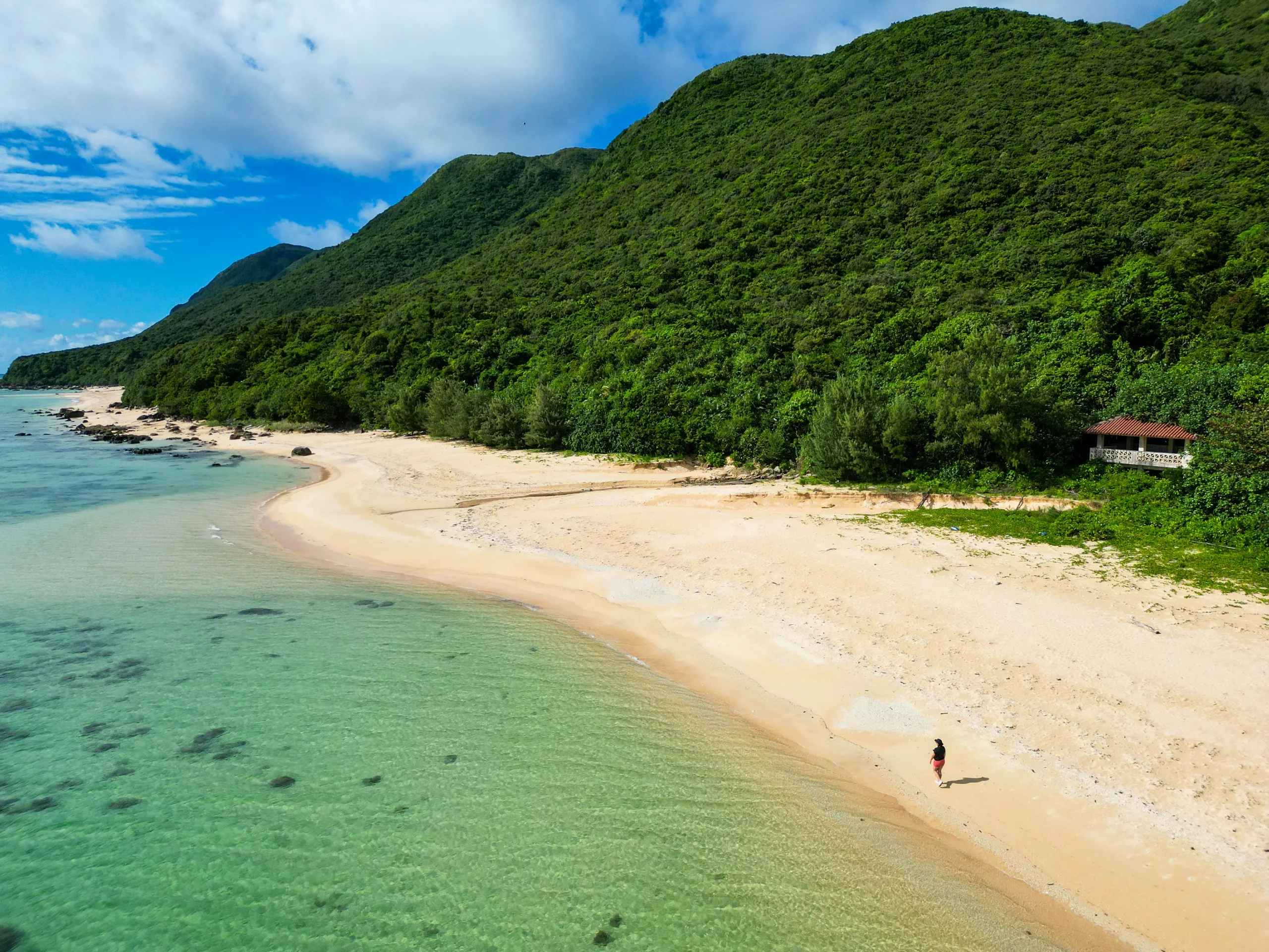 Une personne marche seule sur une plage de sable d'Iriomote, bordée d'eaux turquoise cristallines et de collines verdoyantes et denses, sous un ciel partiellement nuageux. Un petit bâtiment est partiellement visible à droite, près des arbres.