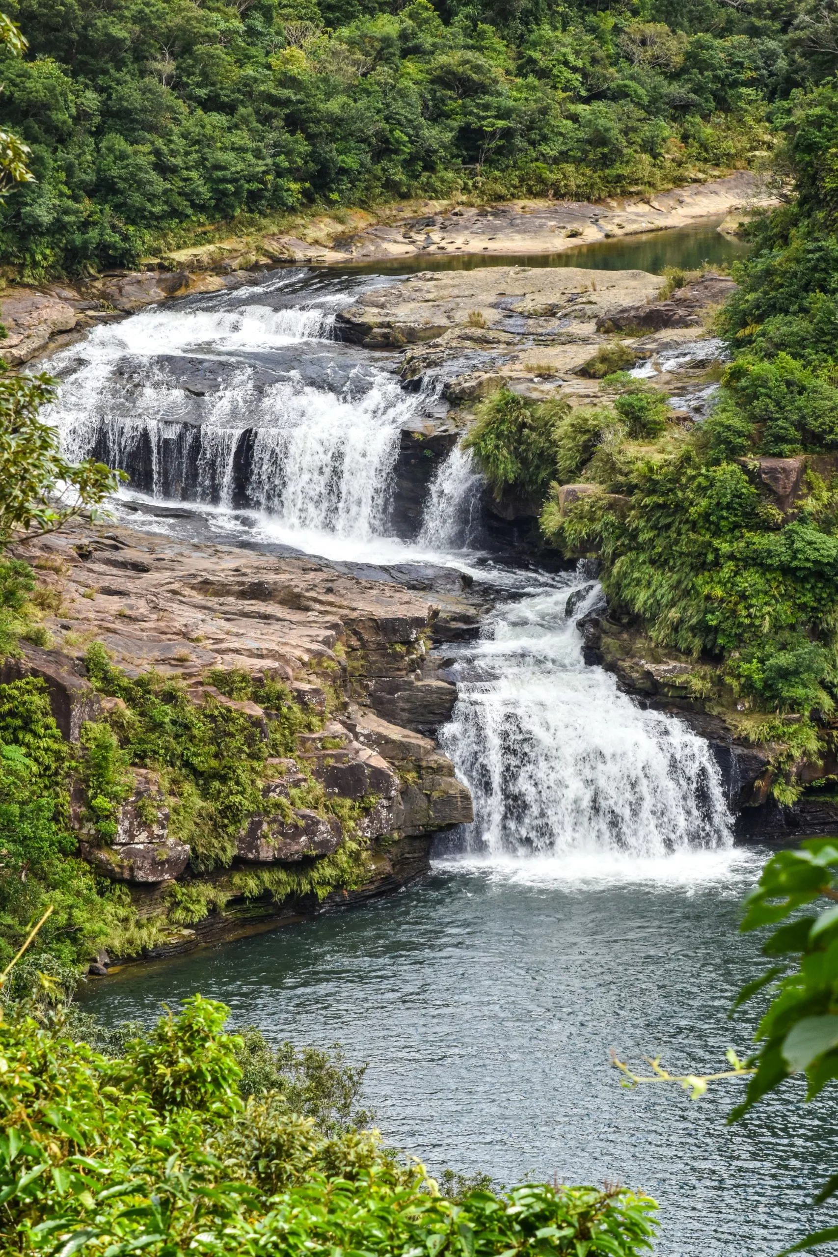 Une cascade à plusieurs paliers dévale des corniches rocheuses entourées d'une végétation dense pour se jeter dans un bassin d'eau calme en contrebas.