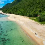 Vue aérienne d'une plage de sable d'Iriomote, avec ses eaux turquoise cristallines, sa forêt verdoyante et dense qui borde le littoral, et une personne marchant au bord de l'eau.