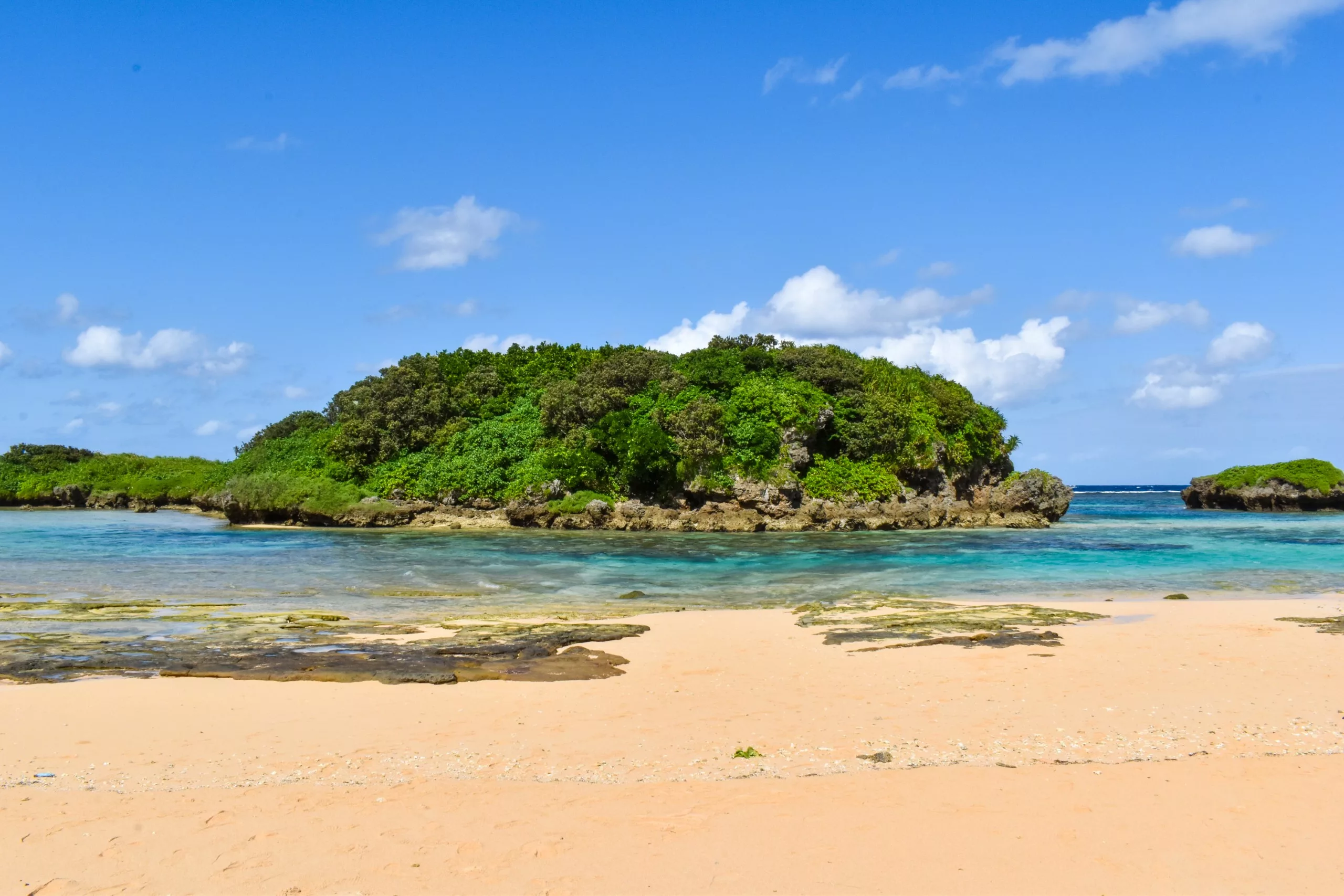 Une petite île verdoyante se détache sur les eaux bleues et limpides, près d'une plage de sable d'Iriomote, sous un ciel radieux parsemé de nuages épars. On aperçoit des rochers et des bassins peu profonds près du rivage.