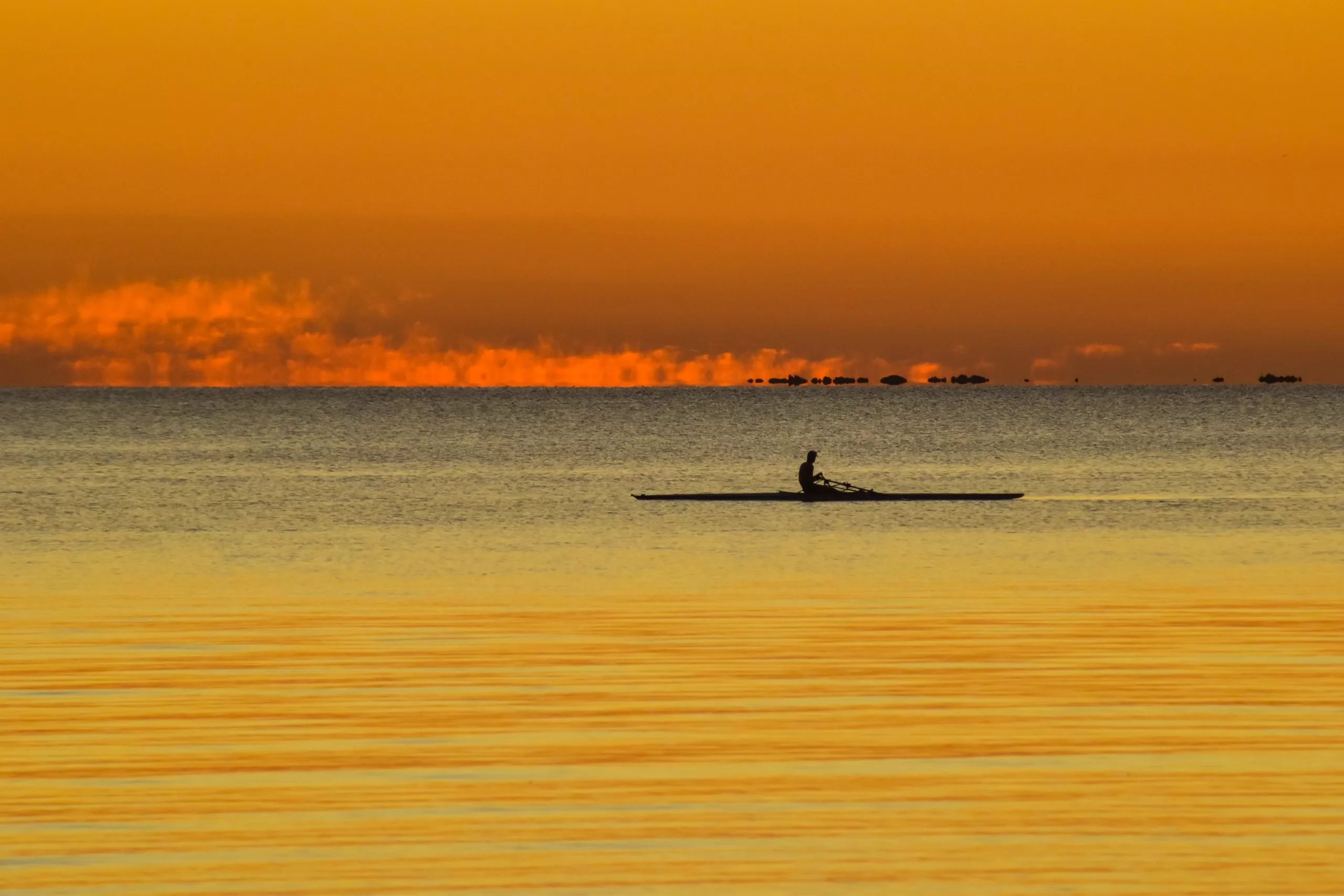 Une personne rame dans un skiff sur des eaux calmes au coucher du soleil, près de Colonia del Sacramento. Le ciel aux teintes orange et jaunes, les nuages bas et cette scène paisible évoquent la paix et la sérénité.