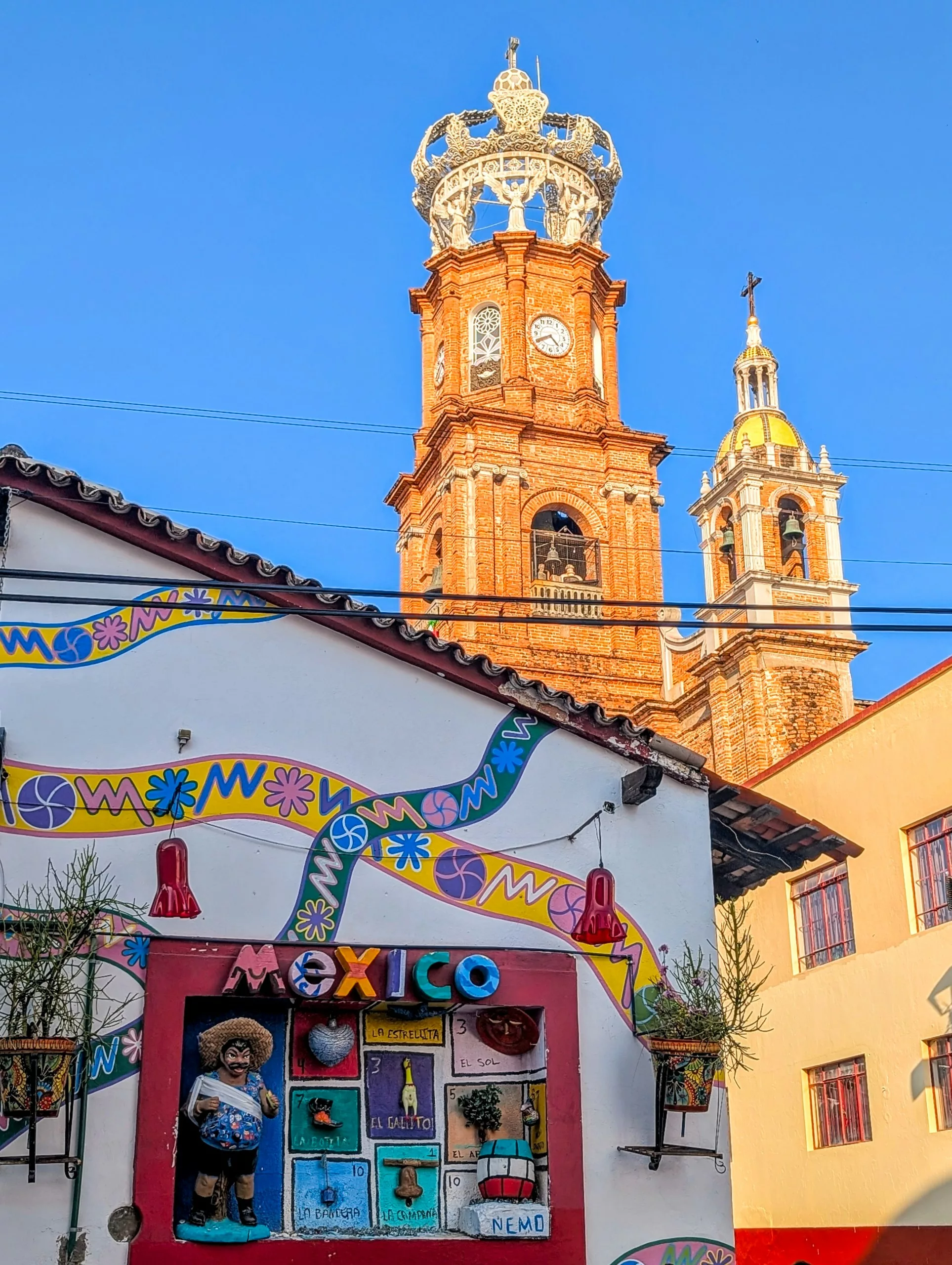 Une devanture décorée, ornée de lettres colorées formant le mot « Mexico », se dresse devant le clocher en briques de l'église de Puerto Vallarta, sous un ciel dégagé.