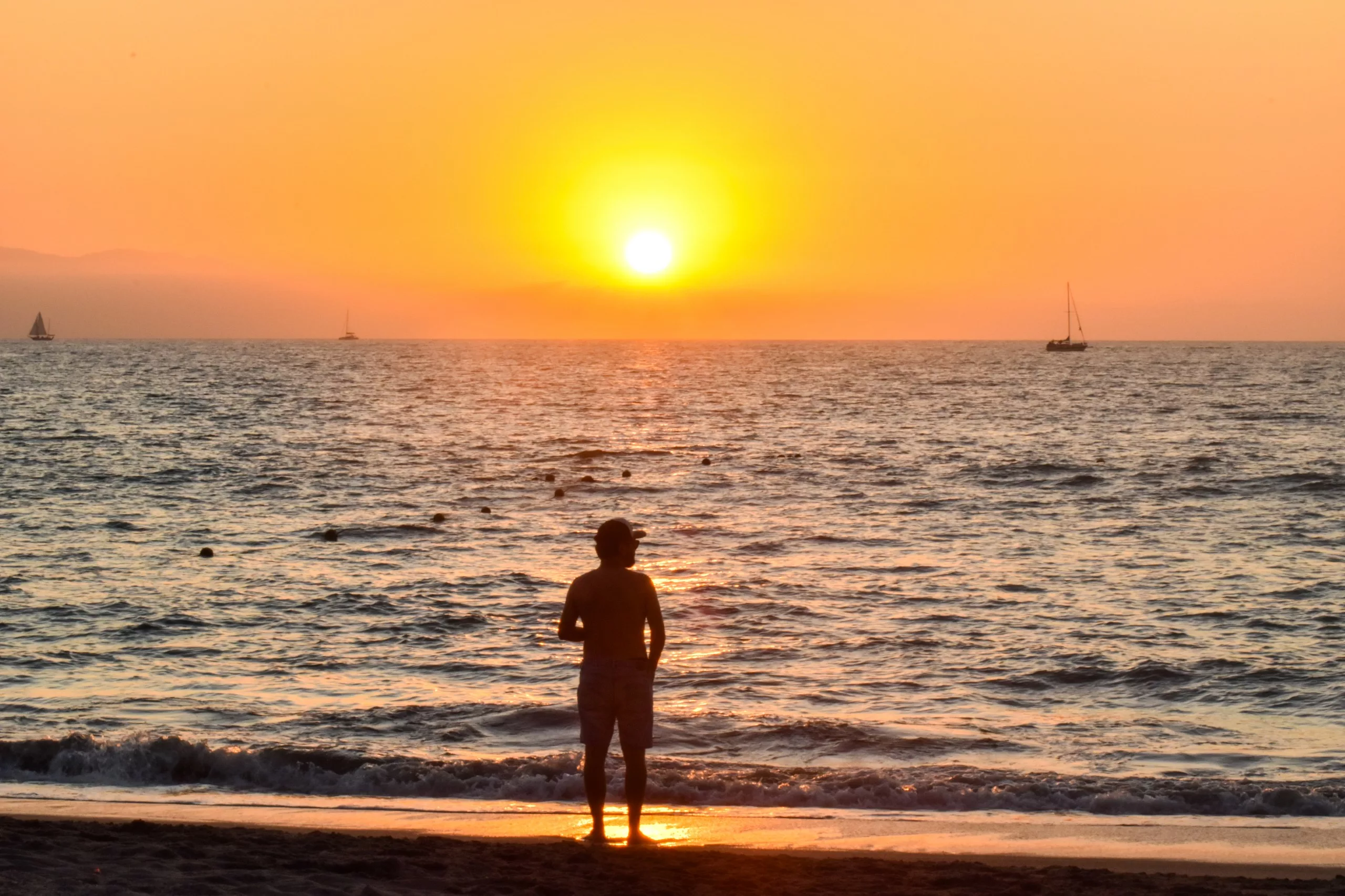 Une personne se tient sur le rivage sablonneux, face à l'océan, au coucher du soleil, et observe deux voiliers : une scène emblématique qui évoque les activités à faire à Puerto Vallarta pour passer une soirée paisible au bord de la mer.