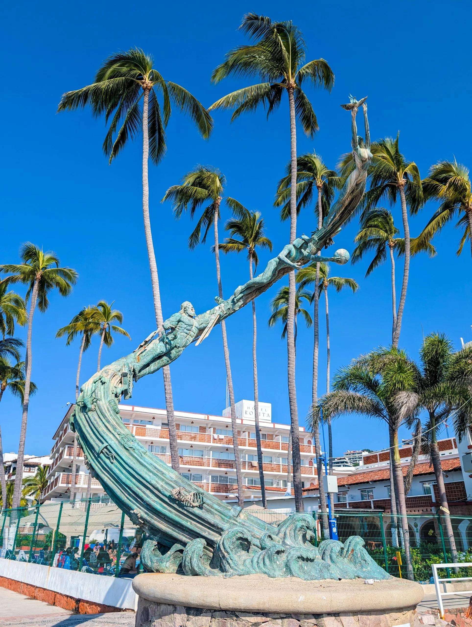 Une sculpture en bronze représentant un homme, une femme et des dauphins émerge de vagues stylisées encadrée par des palmiers et un ciel bleu.