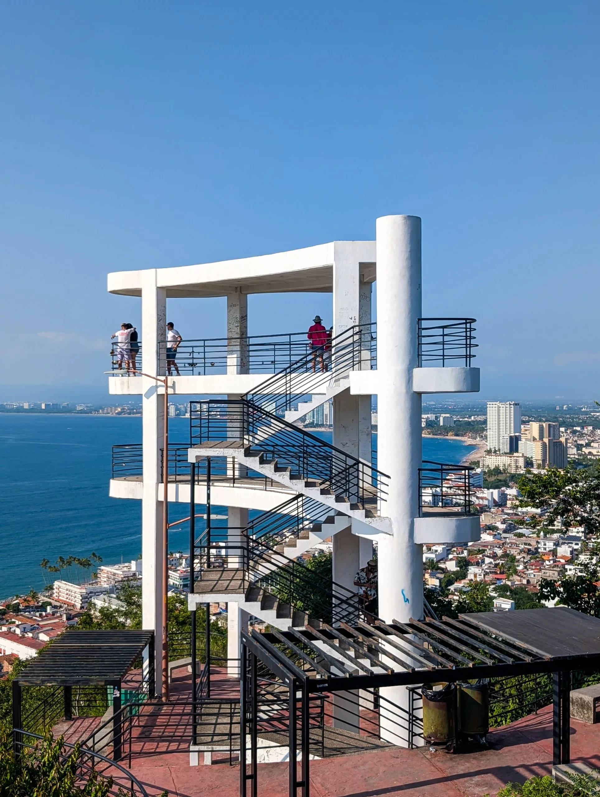 Une grande tour d'observation blanche et moderne, dotée d'un escalier, se dresse sur une colline à Puerto Vallarta et offre une vue panoramique sur la côte sous un ciel bleu limpide.