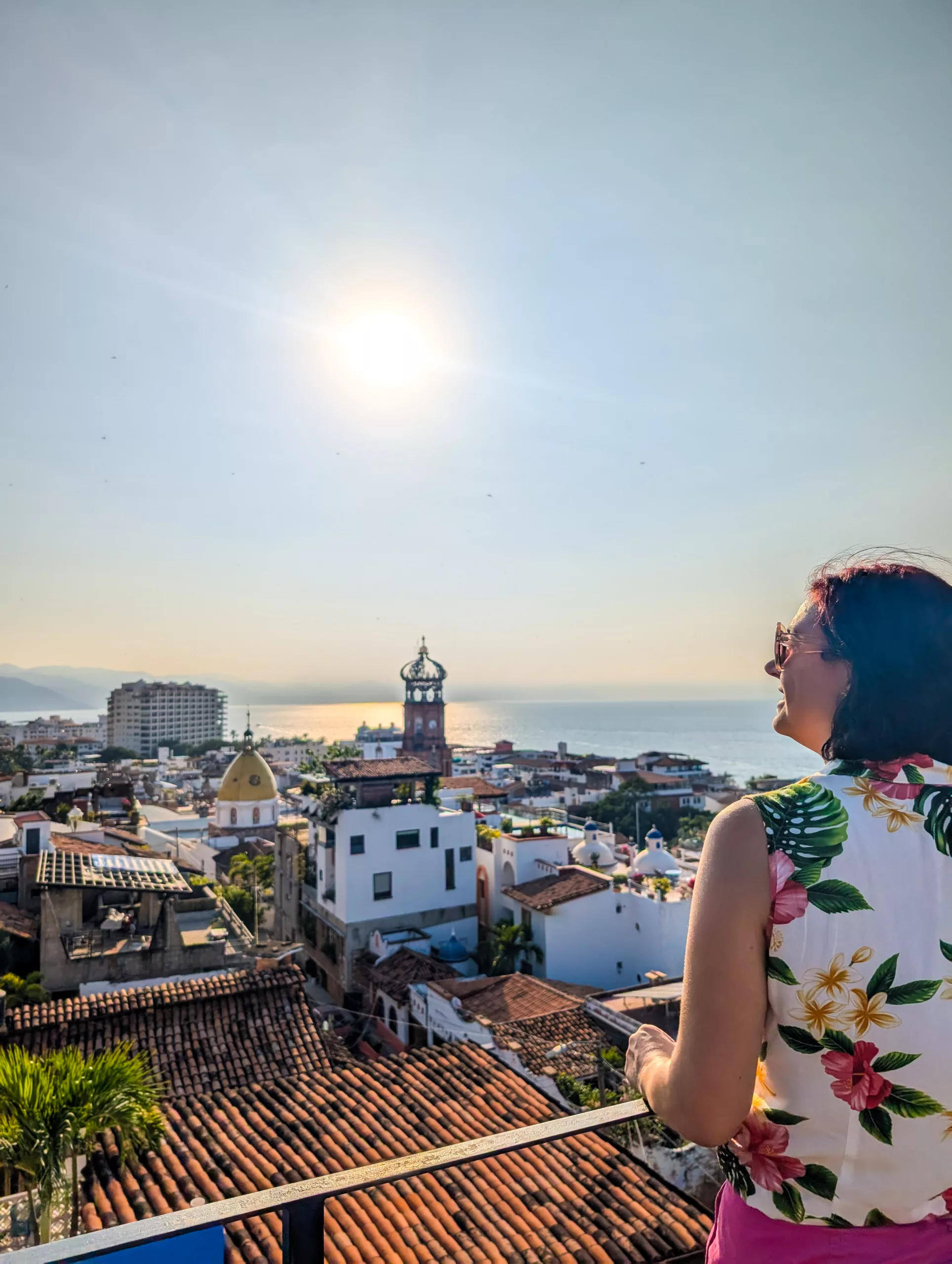 Une femme vêtue d'une chemise à fleurs se tient sur un toit, face à Puerto Vallarta, un paysage urbain côtier baigné de soleil avec ses toits de tuiles rouges, sa tour et ses superbes vues sur l'océan.
