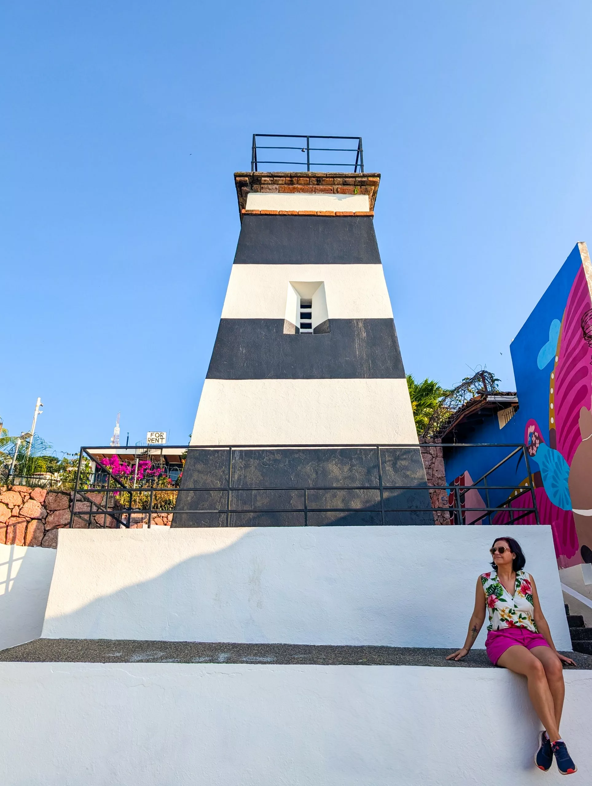 Une femme portant des lunettes de soleil est assise sur un mur blanc devant un phare rayé de noir et blanc ; à sa droite, une fresque murale colorée ; au-dessus d'elle.