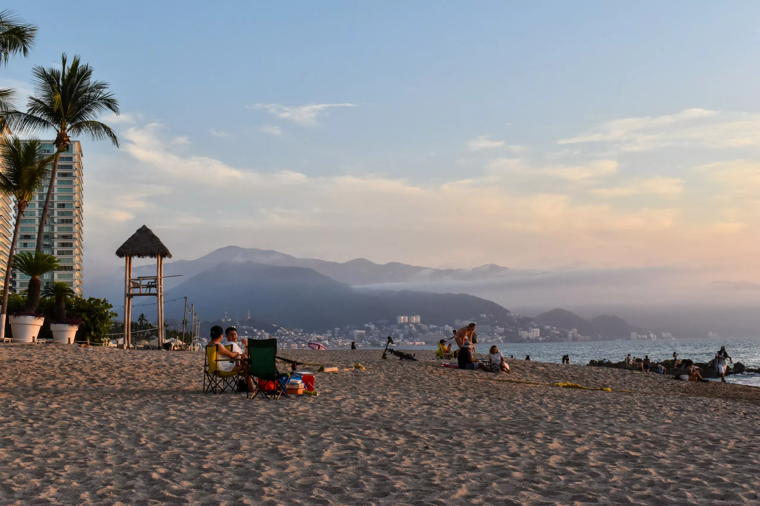Des personnes sont assises sur une plage de sable bordée de palmiers à Puerto Vallarta, avec un poste de sauveteurs et de grands immeubles à proximité, au coucher du soleil