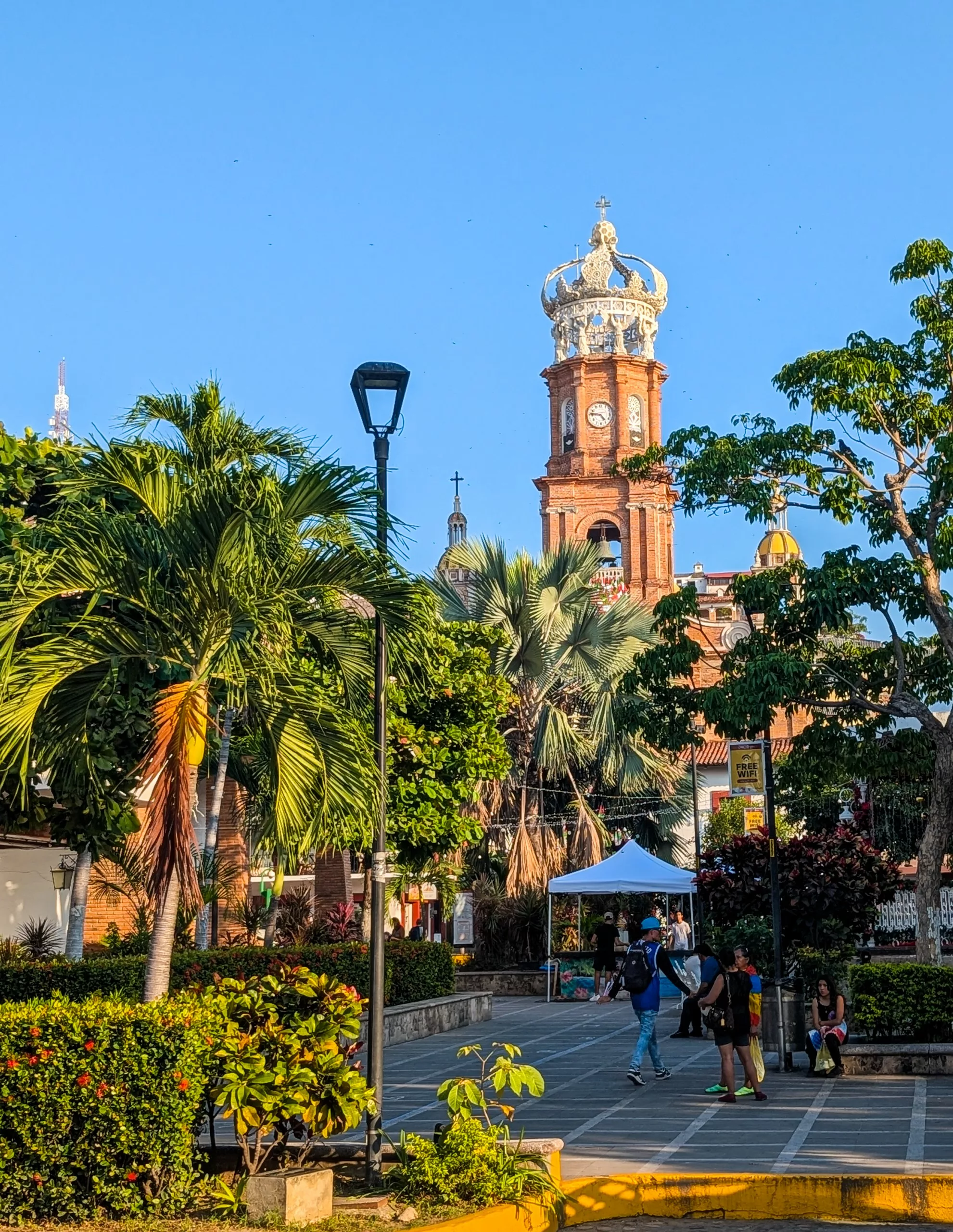 Des gens se promènent et s'assoient sur une place agrémentée de plantes tropicales. À l'arrière-plan se dresse une église en briques dotée d'une structure en forme de couronne et d'une tour horloge.