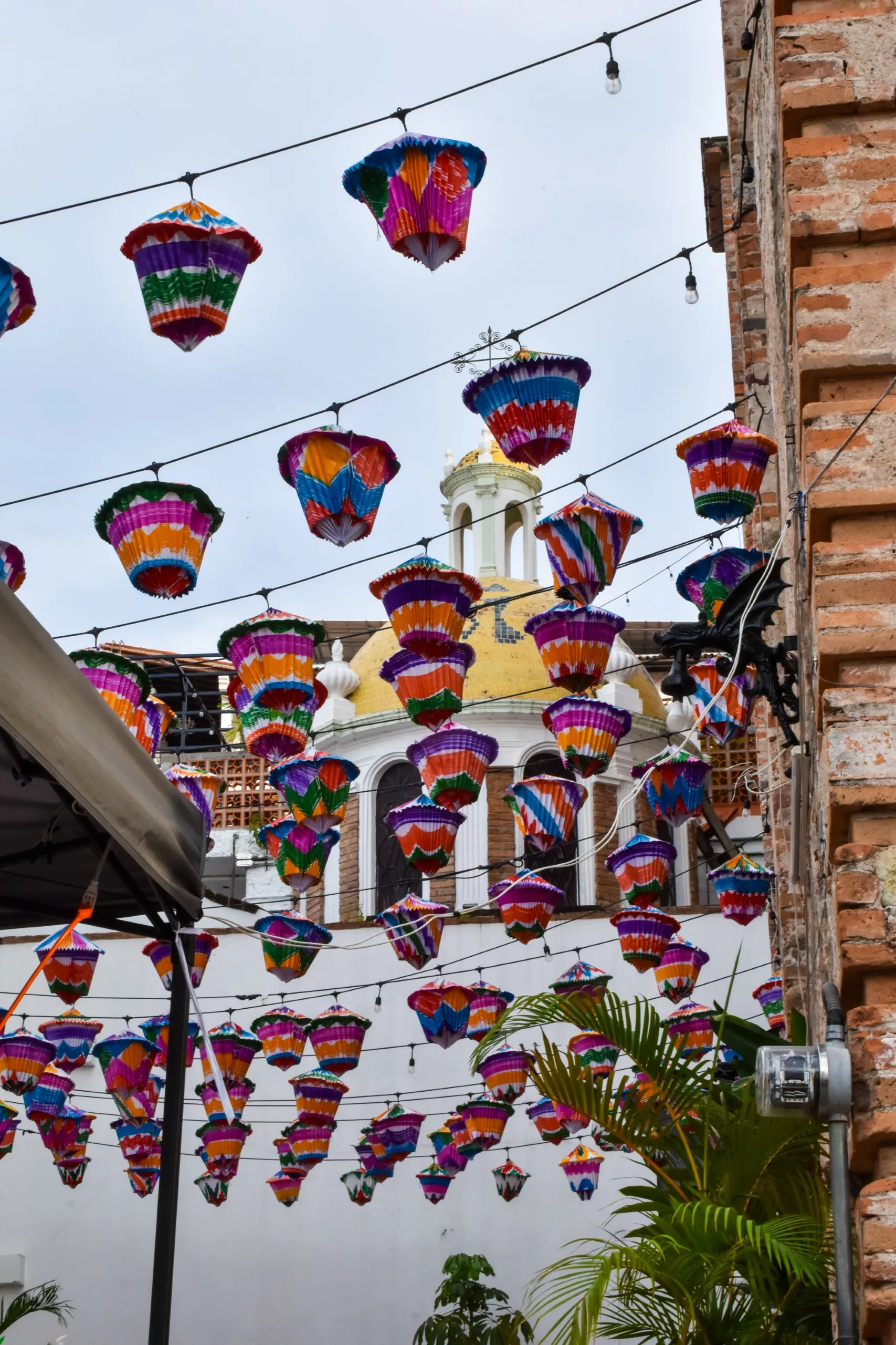 Des lanternes en papier aux couleurs vives sont suspendues au-dessus d'un espace extérieur situé entre des bâtiments en briques, avec de la verdure, un auvent de tente et une église au dôme jaune en arrière-plan