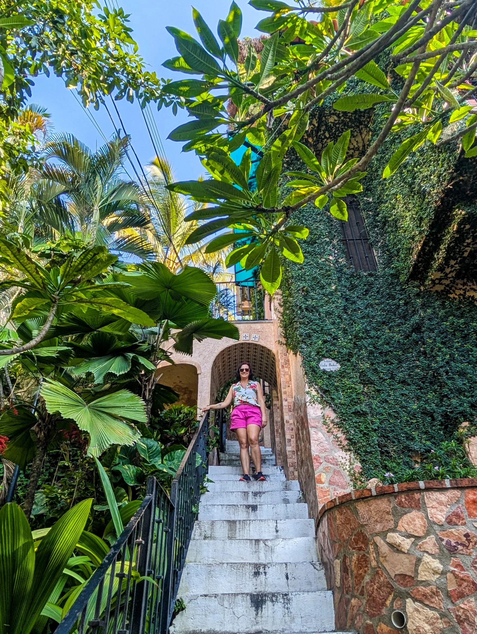 Une personne portant des lunettes de soleil et un short rose se tient sur un escalier blanc, entourée d'une végétation luxuriante