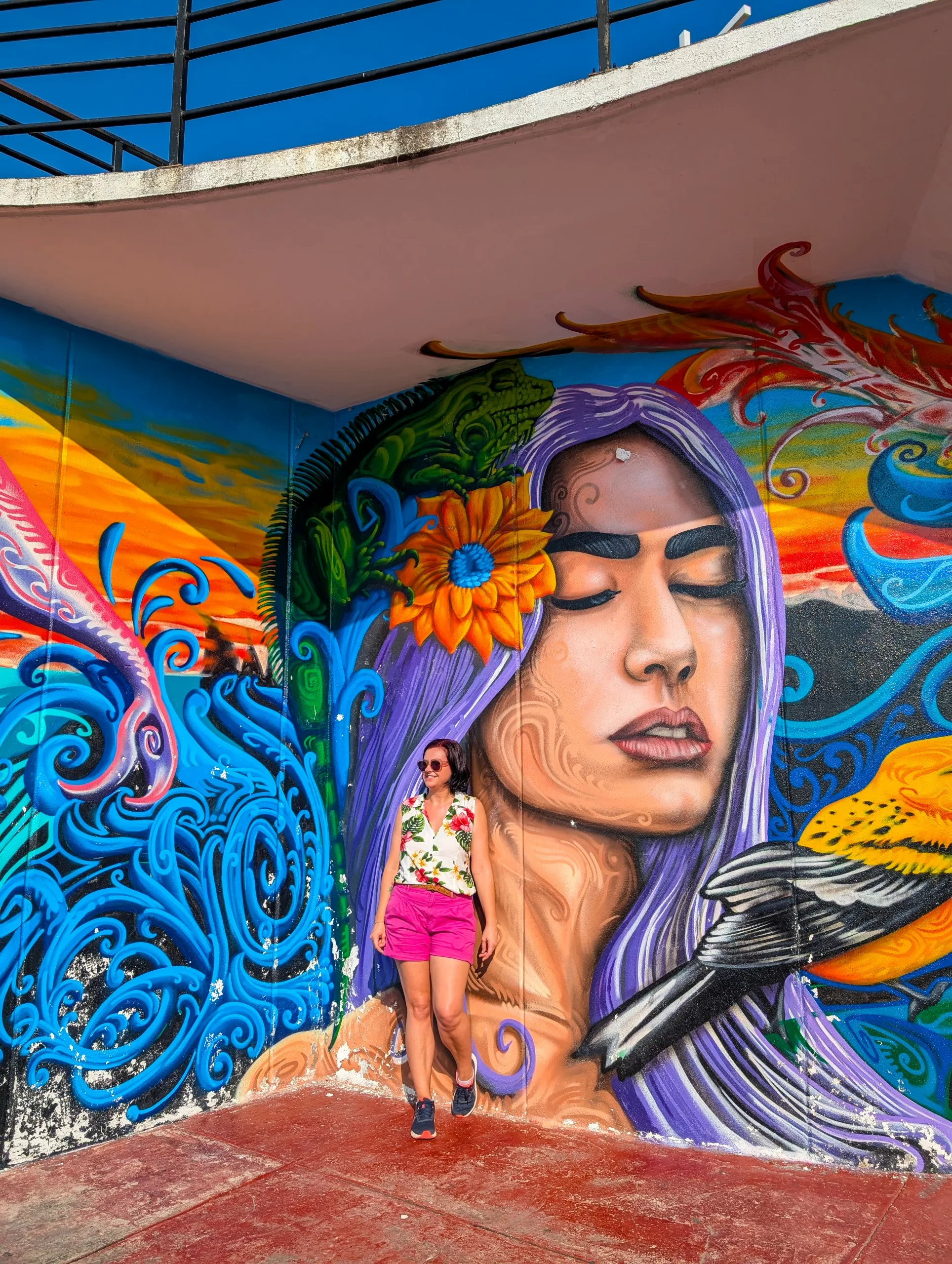 Une personne portant des lunettes de soleil, un haut à fleurs et un short rose se tient devant une fresque murale colorée représentant une femme aux cheveux violets à Puerto Vallarta