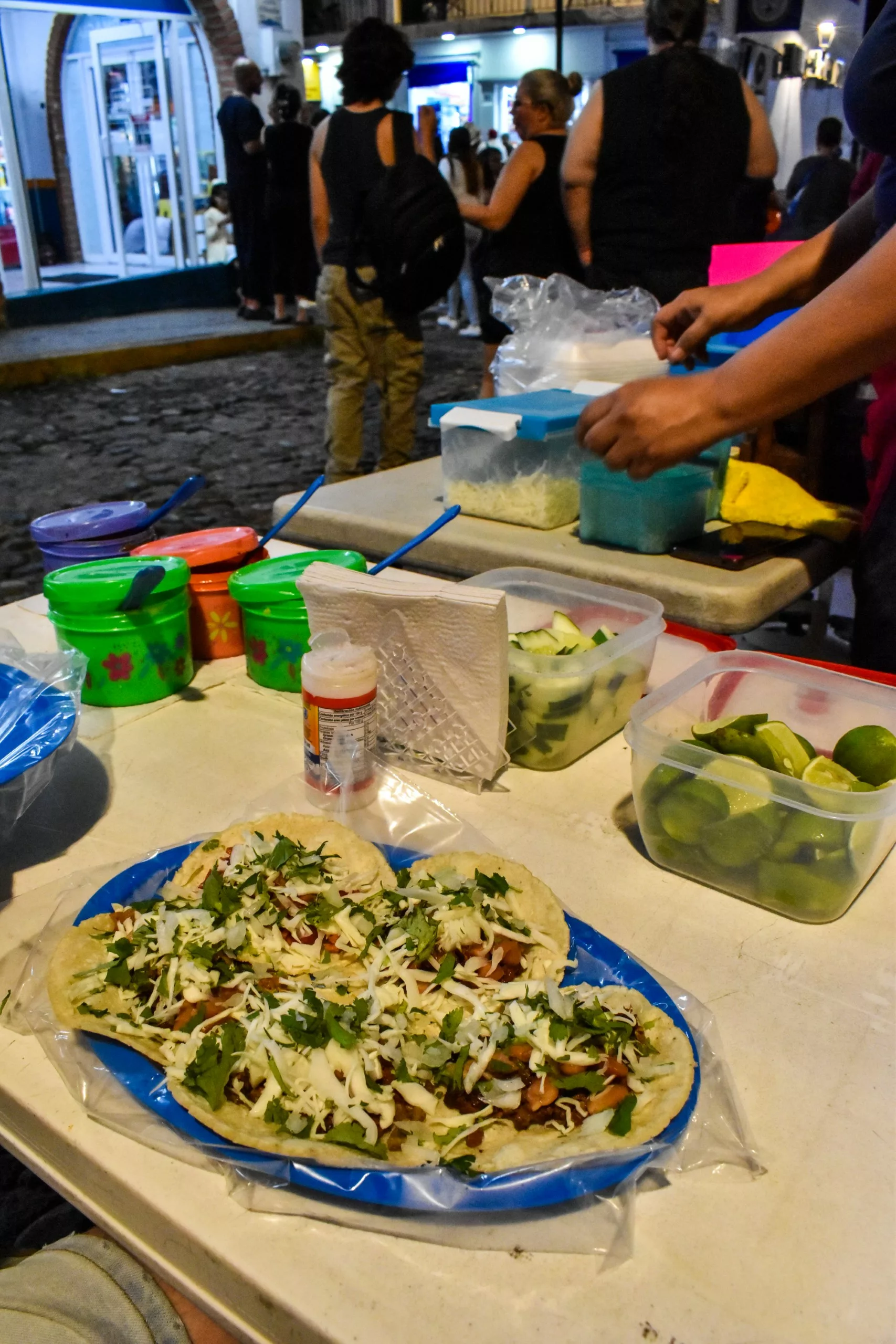 Une assiette de tacos garnis d'oignons et de coriandre trône sur une table où sont disposés des citrons verts, des sauces et des condiments.