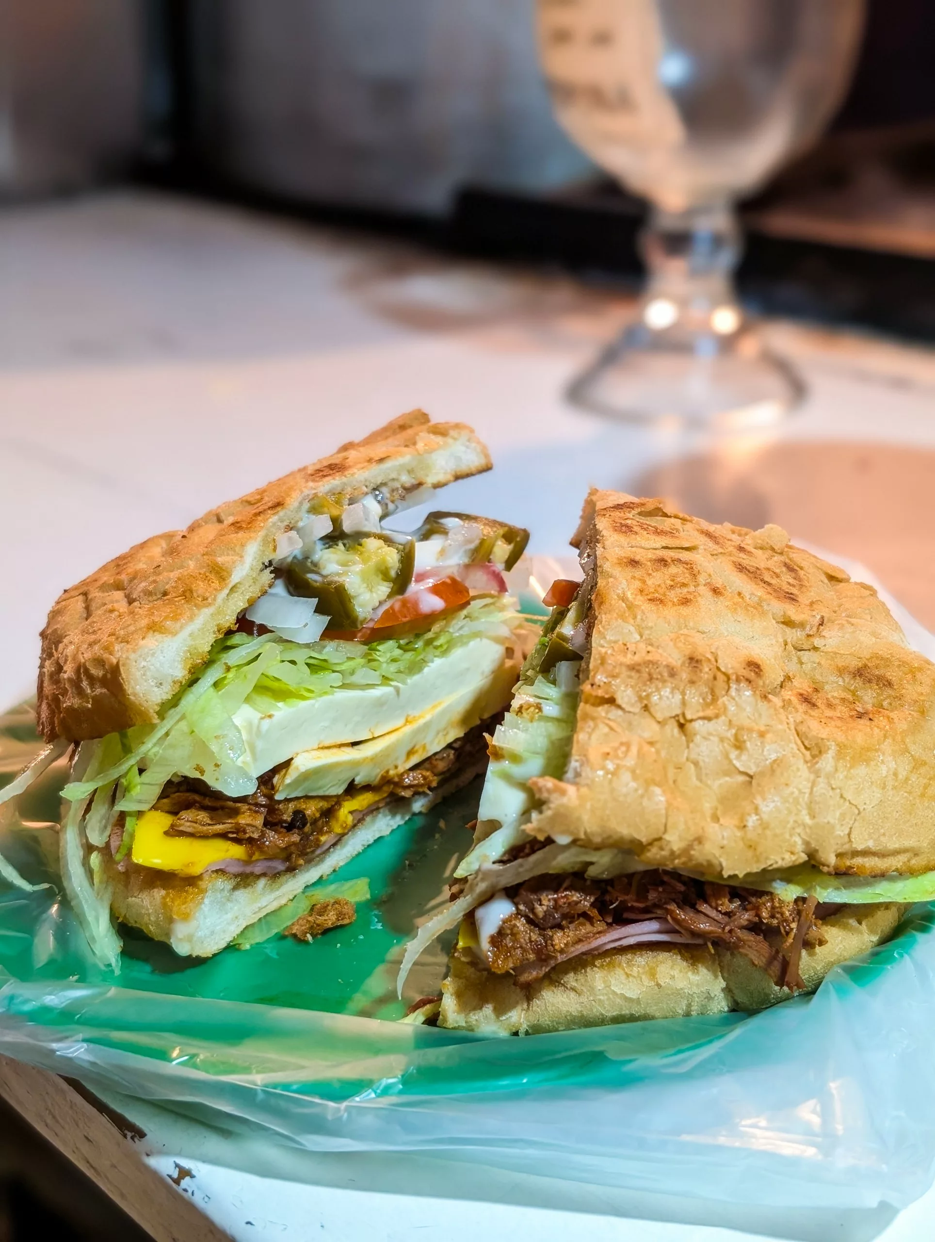 Un sandwich coupé en deux repose sur une nappe en plastique verte. Il est garni de viande effilochée, de laitue, de tomate, d'oignons, de fromage et de piment, le tout dans un petit pain croustillant.