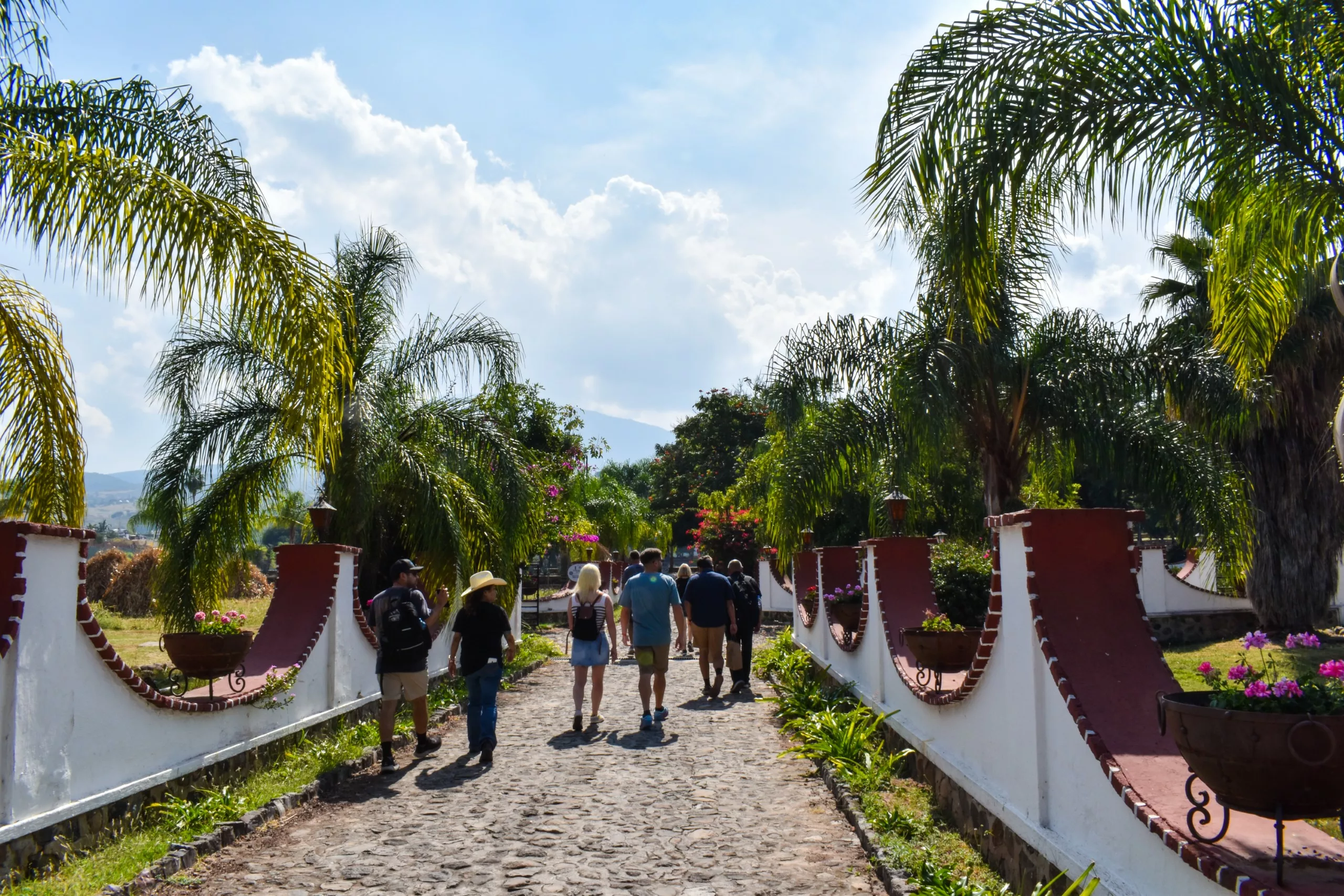 Un groupe de personnes se promène sur une allée pavée dans la ville de Tequila, au Mexique, bordée de murs blancs et rouges et de palmiers, sous un ciel partiellement nuageux.