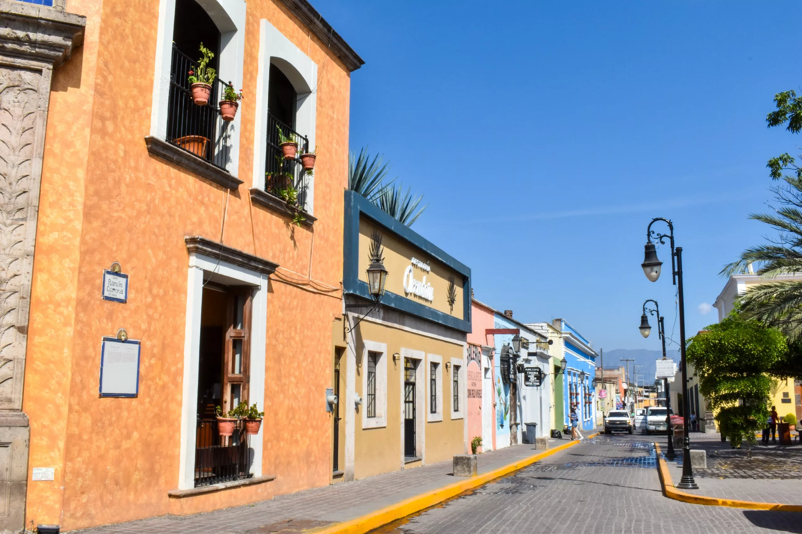 Des bâtiments colorés de style colonial bordent une rue pavée de la ville de Tequila, sous un ciel bleu limpide. Des plantes en pot ornent les fenêtres, un lampadaire se dresse à proximité et une voiture blanche est garée au loin.