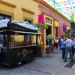 Une scène de rue animée dans la ville de Tequila, au Mexique, où les gens se pressent près d'un chariot noir de tequila « Jose Cuervo Express », sous des décorations festives en papel picado et devant des bâtiments aux couleurs vives.